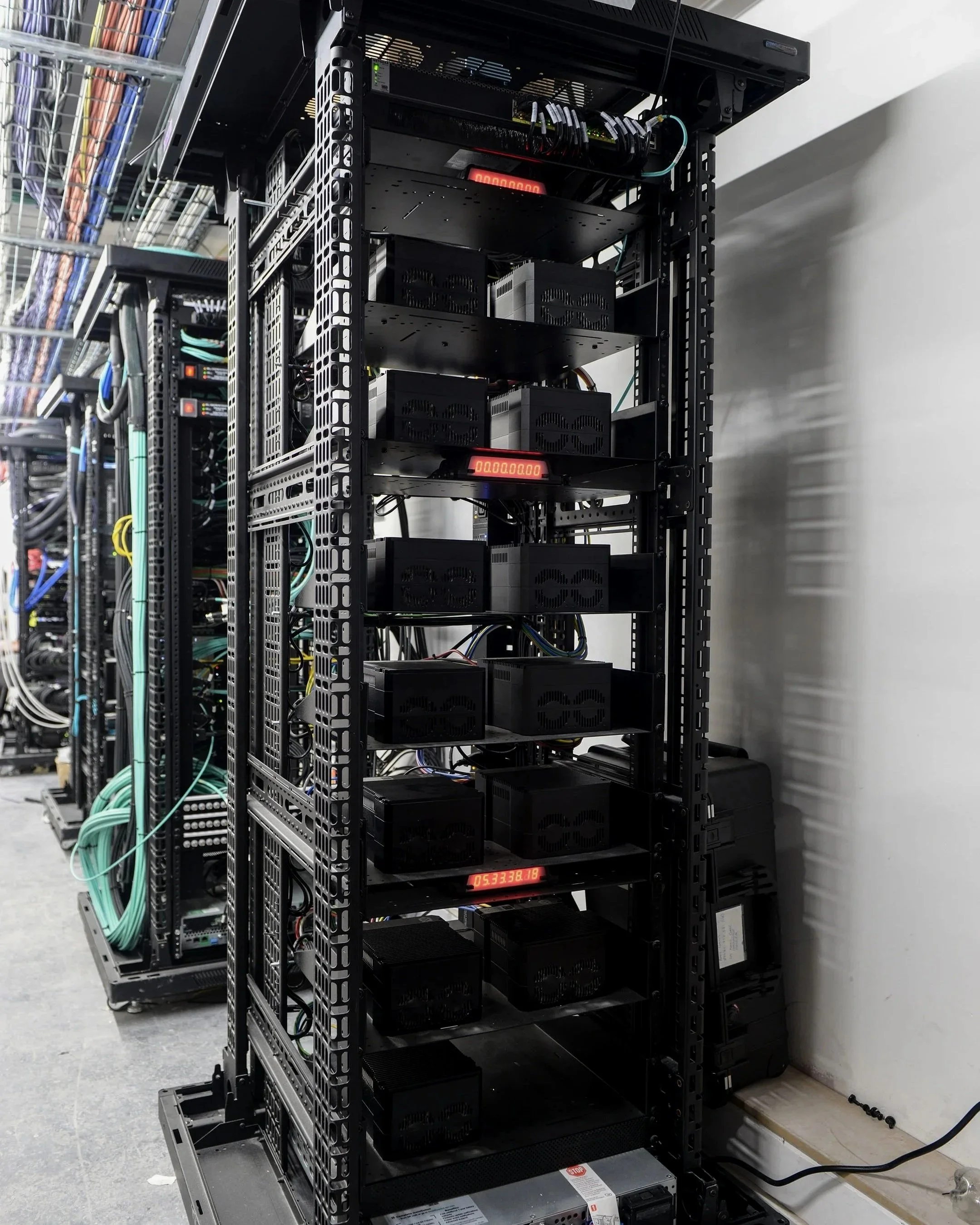 A server rack filled with multiple black power supplies, and electronic components, with digital displays showing numbers, situated in a data center or server room with cables connected to the equipment.