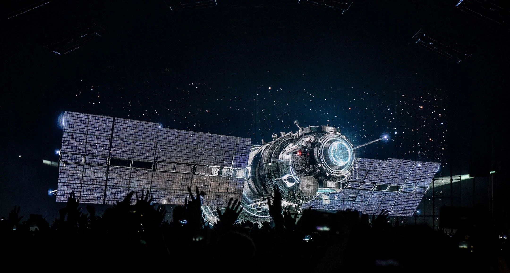 A spacecraft with solar panels is shown in space against a starry background, with a crowd of people watching in silhouette in the foreground.