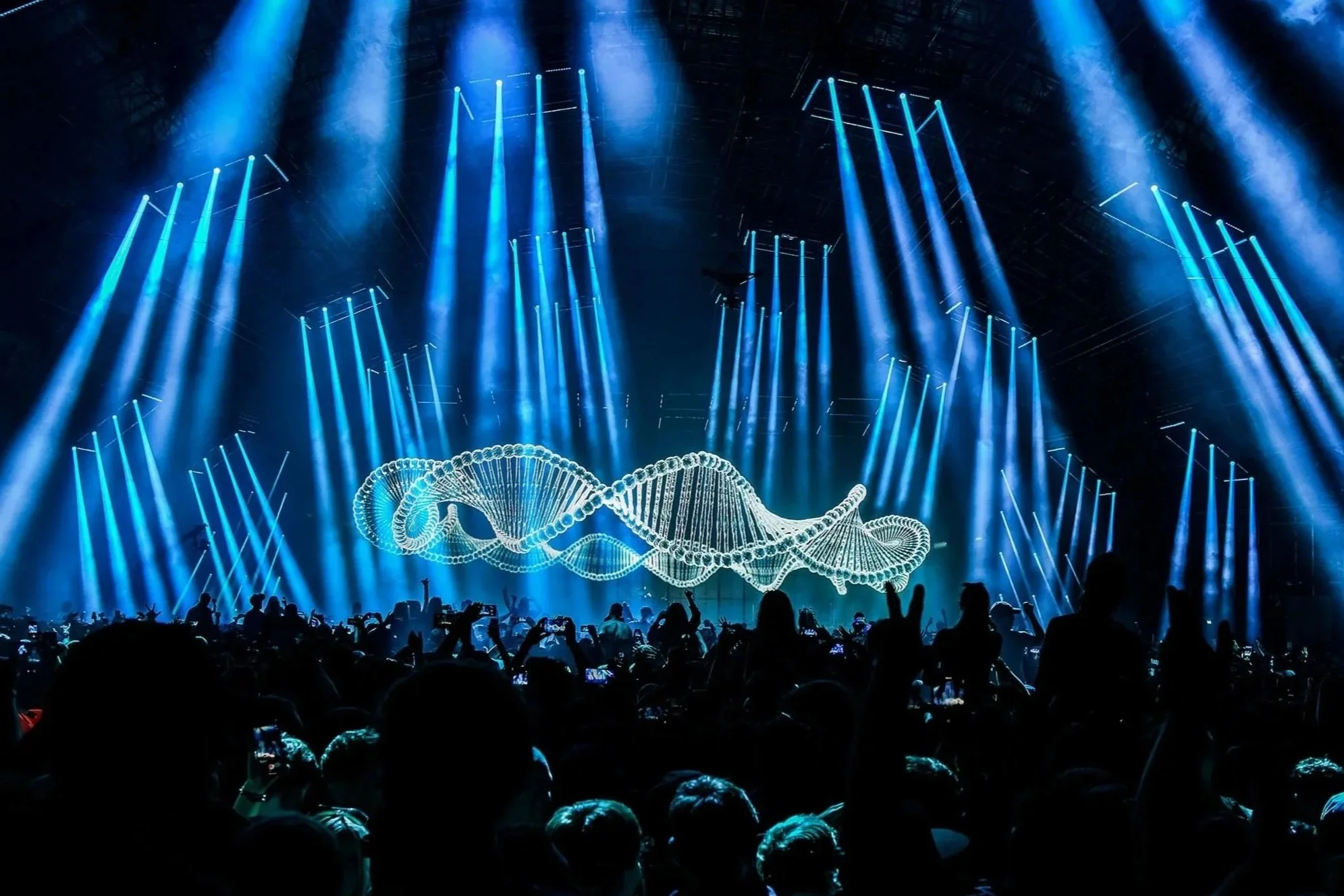 A concert stage with bright blue lights and a large illuminated DNA double helix projection in the background, and a crowd of people watching.