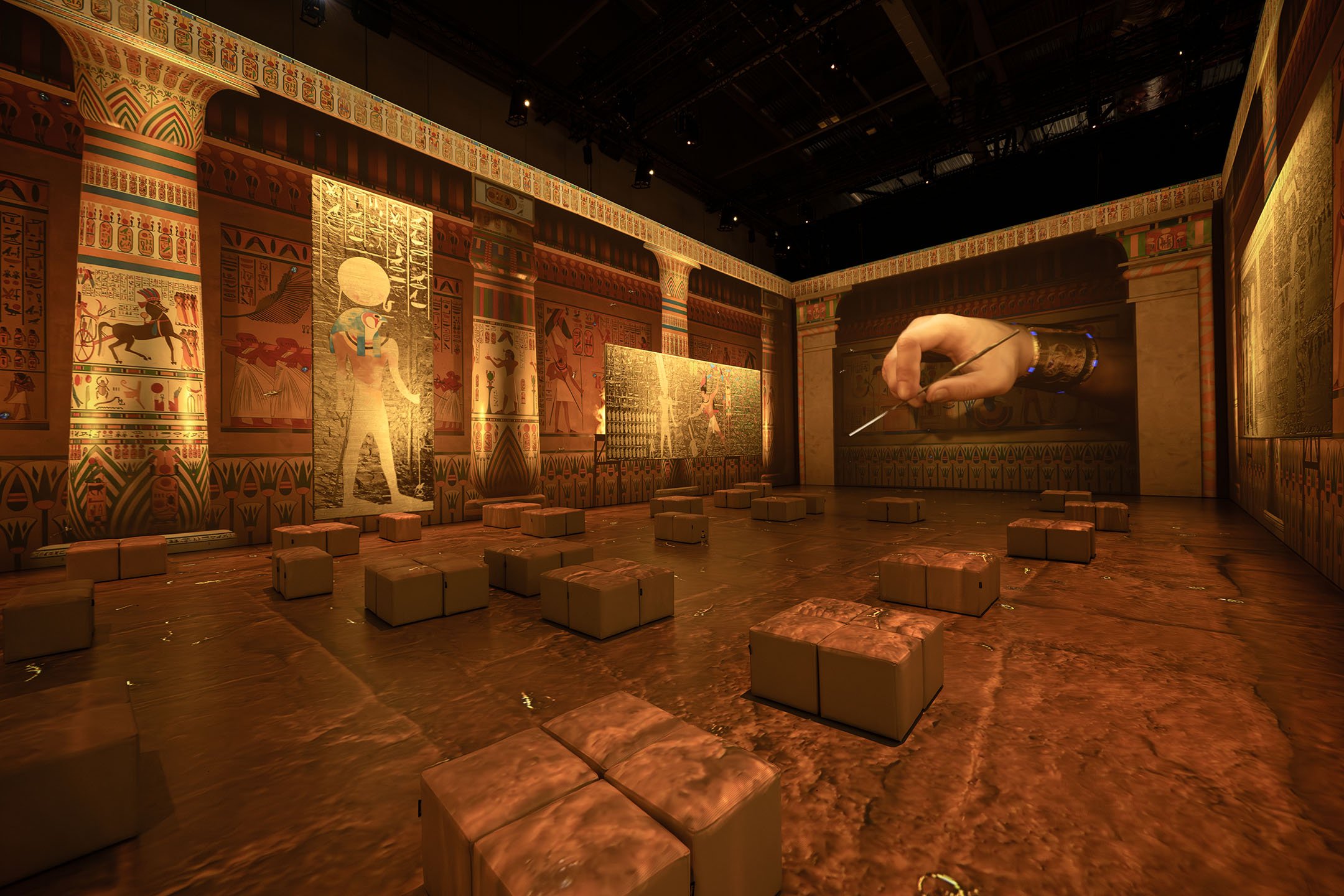 Museum exhibit with Egyptian-themed wall art, a large floating hand with a pointing finger, and seating cubes on a theater-like stage.