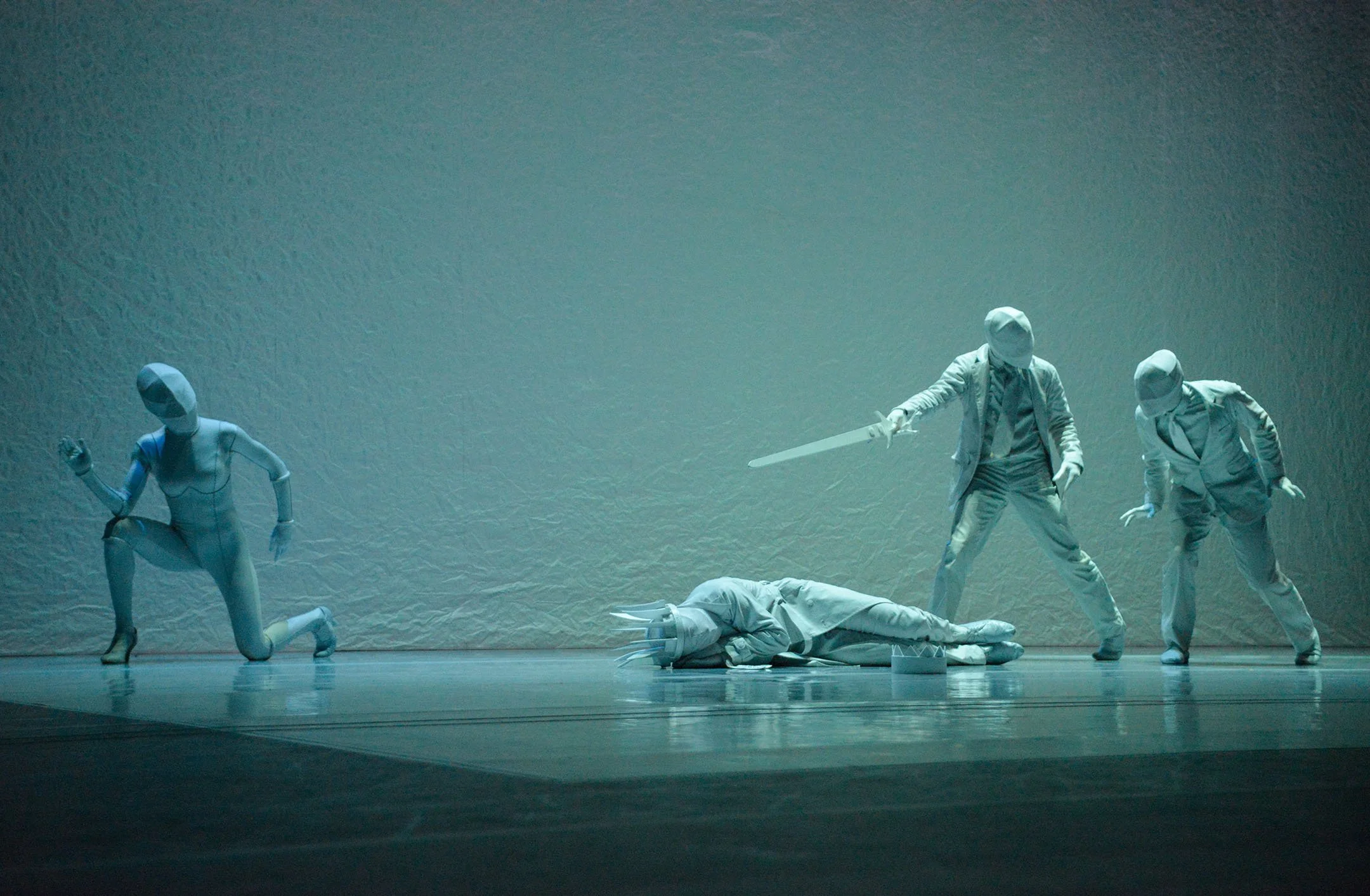Stage scene with four white humanoid figures, one kneeling, one lying down, and two standing, one holding a sword, in a minimalist, textured background.