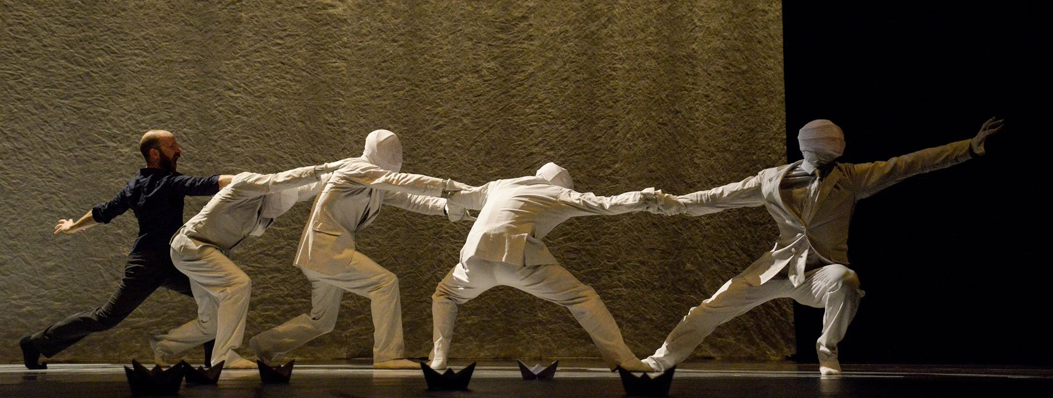 A stage performance depicting a man and four figures dressed in white, with three of them with their faces covered with cloth, all reaching forward in a synchronized dance move against a textured brown and black background.