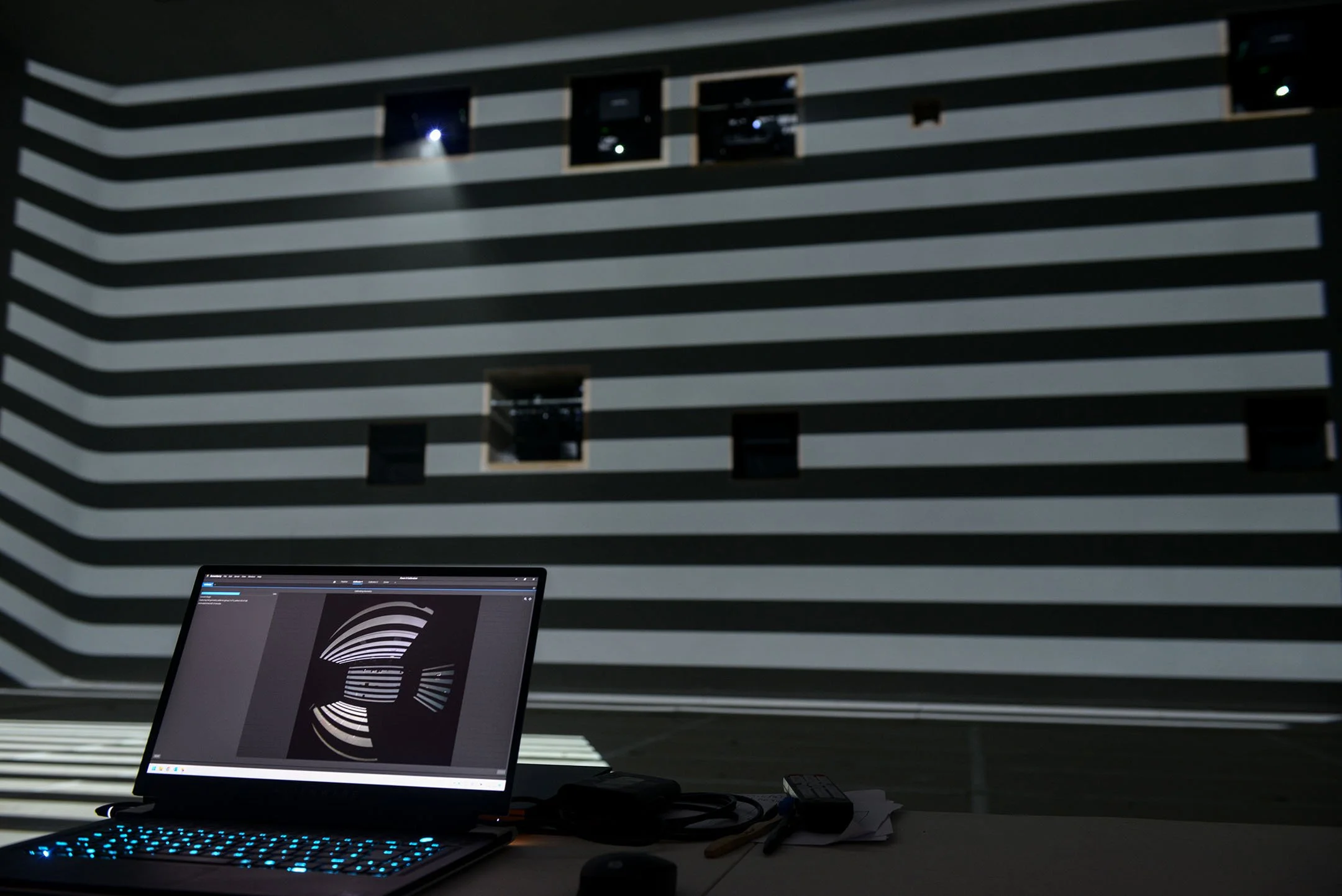 A laptop displaying a projection of an optical illusion pattern of black and white stripes on its screen, set up on a desk with various electronic devices and papers in front of a wall with horizontal black and white striped pattern.