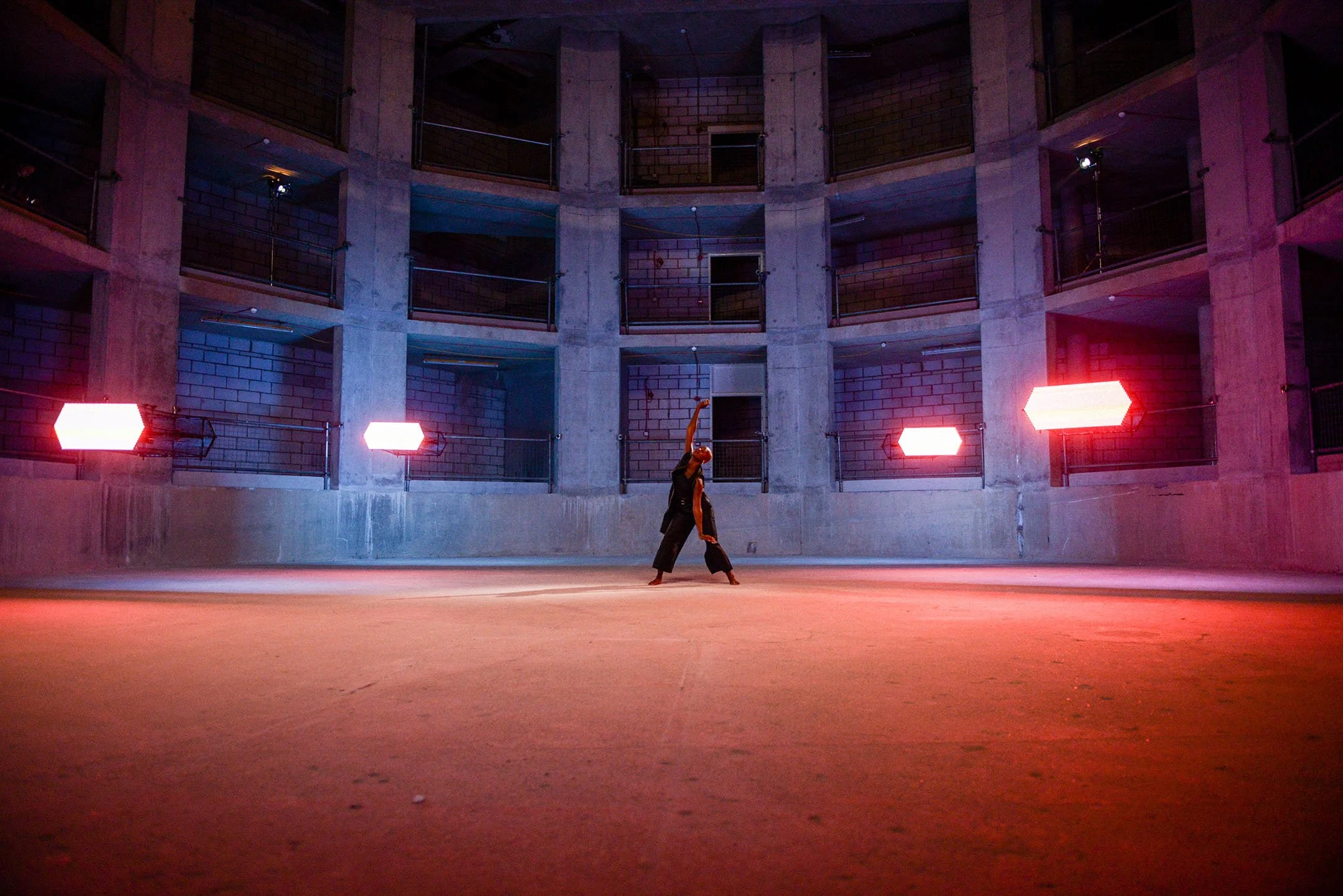 A dancer performing in an empty, circular concrete room with multiple levels of balconies. The space is lit with pink and red neon lights.