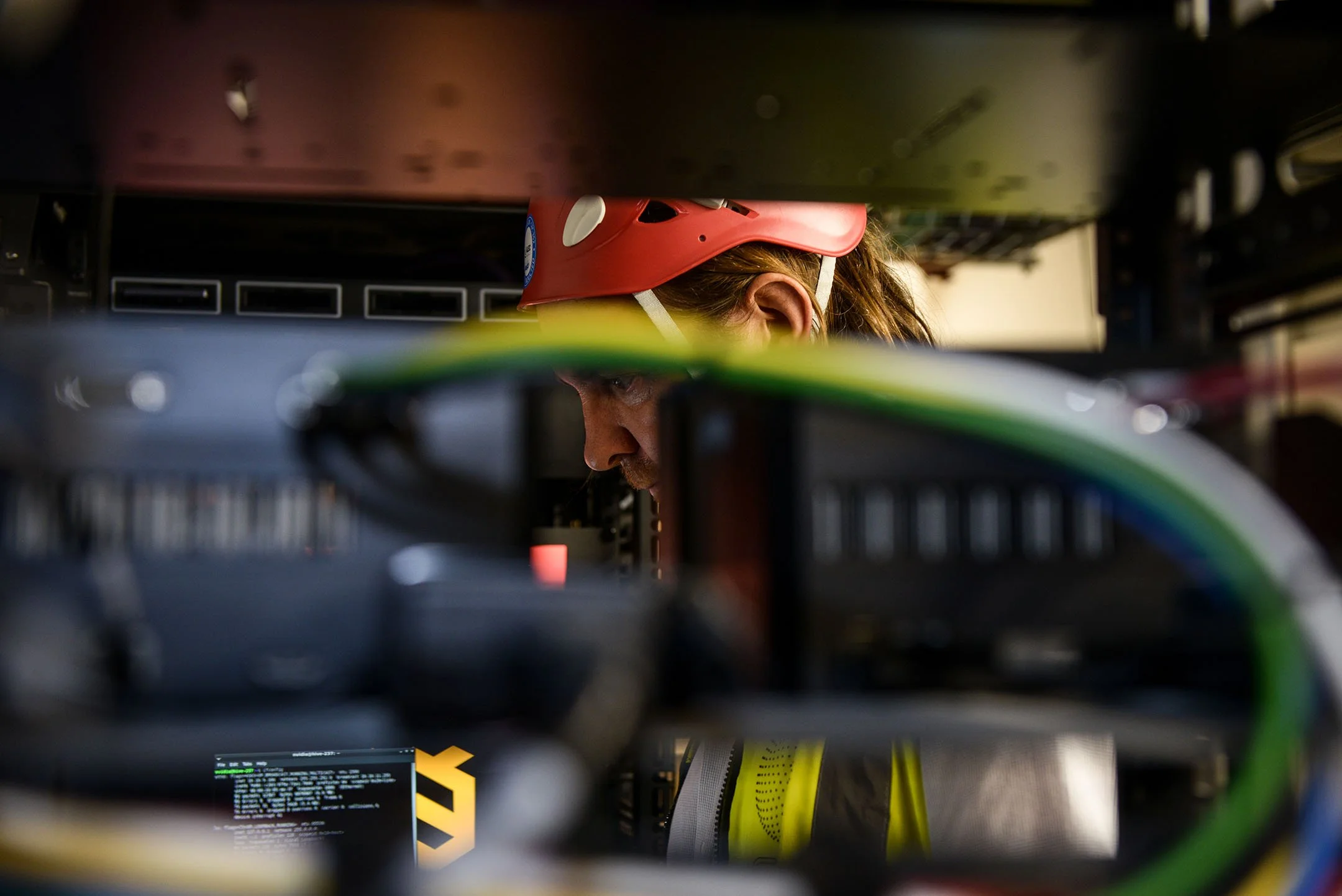 A woman with a red hard hat working on computer hardware, seen through computer components and wires.