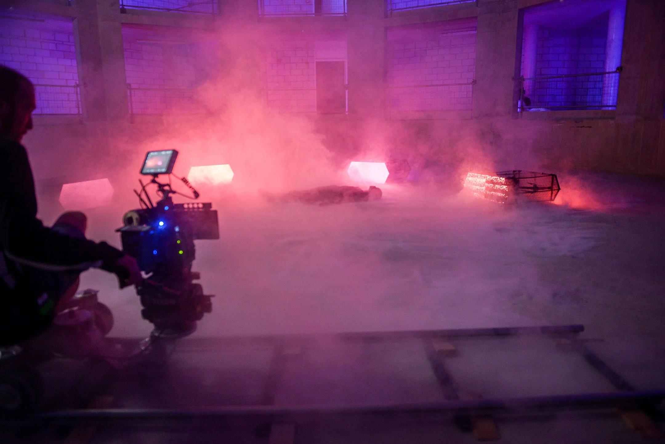 Behind-the-scenes shot of a movie or TV production showing a person operating a camera on a dolly, with fog and bright colored lights in a large indoor studio space.