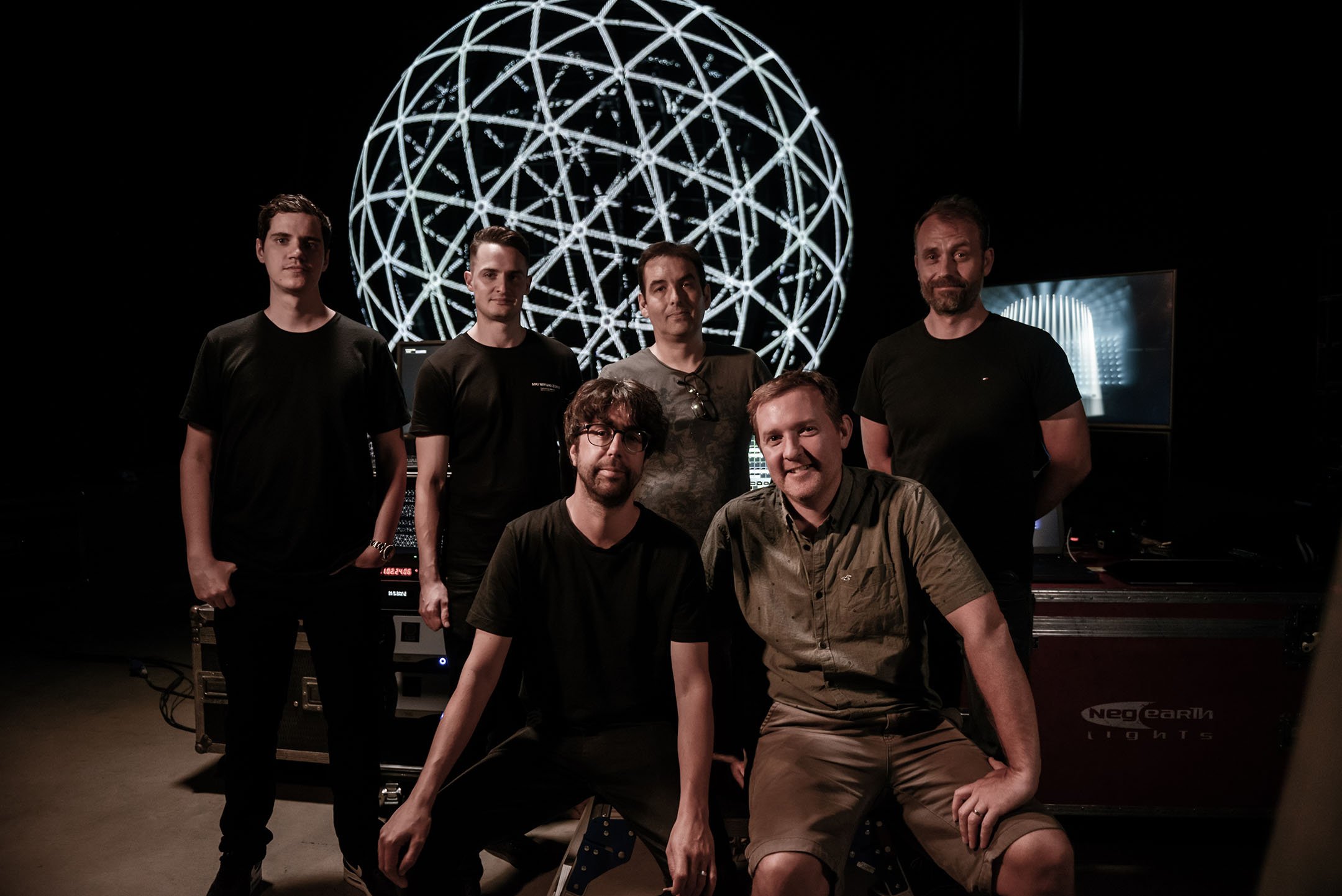 Group of six men in a dark room with electronic equipment and a large illuminated geometric globe in the background.