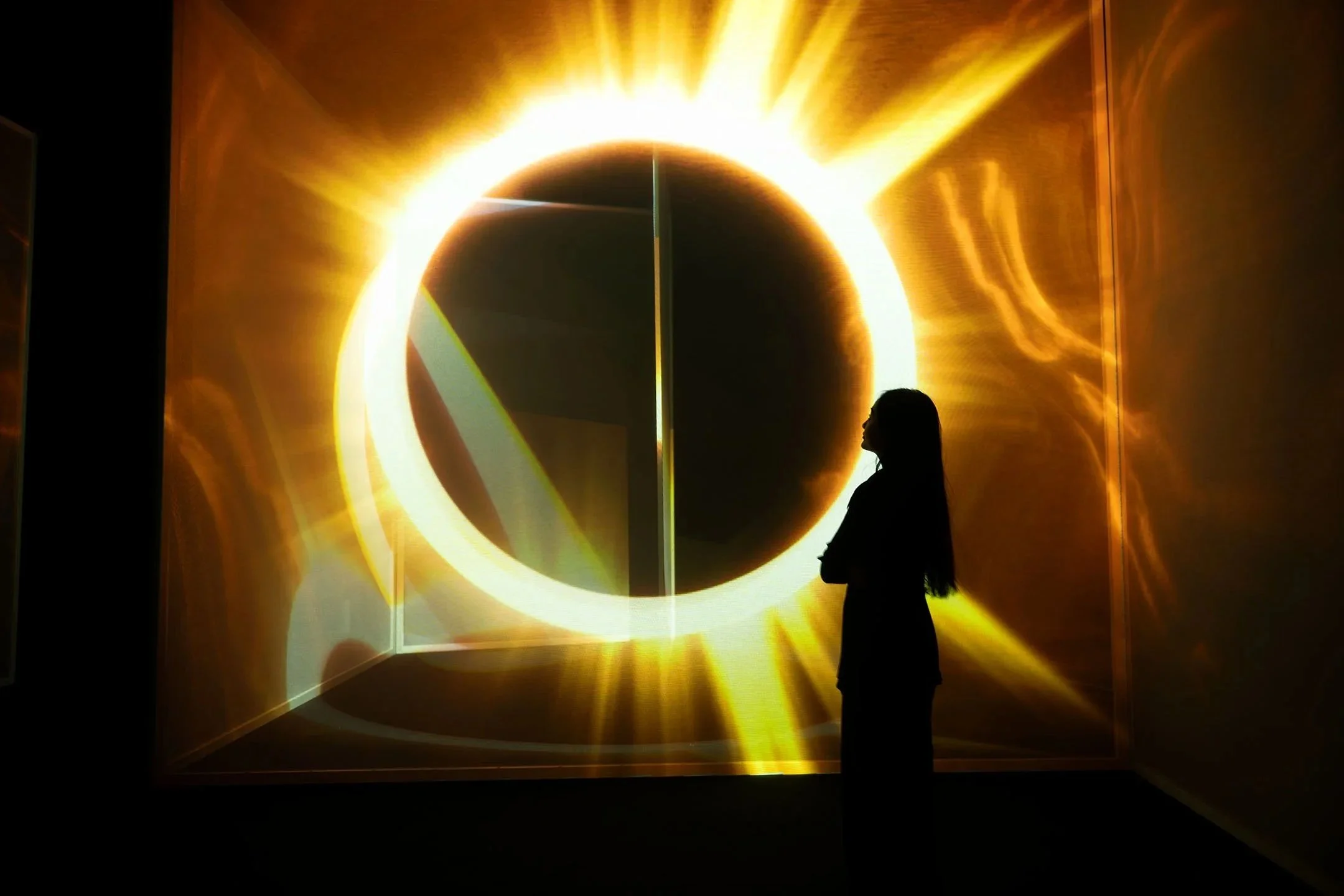 A person standing in front of a large digital display of a solar eclipse with a bright ring of sunlight and rays emanating outward.