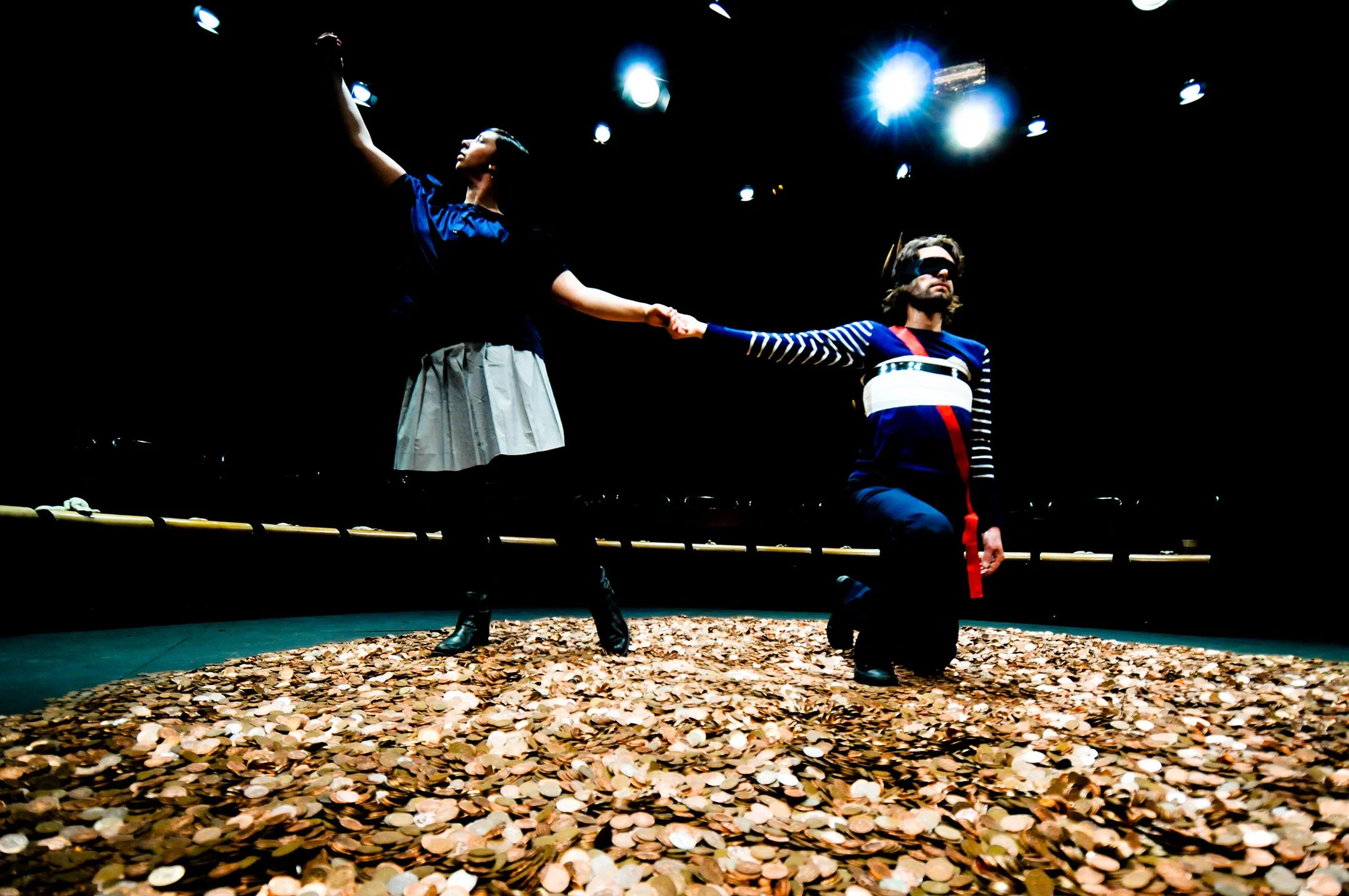 Two performers on stage, one kneeling and the other standing, holding hands, surrounded by coins on the floor under stage lights.