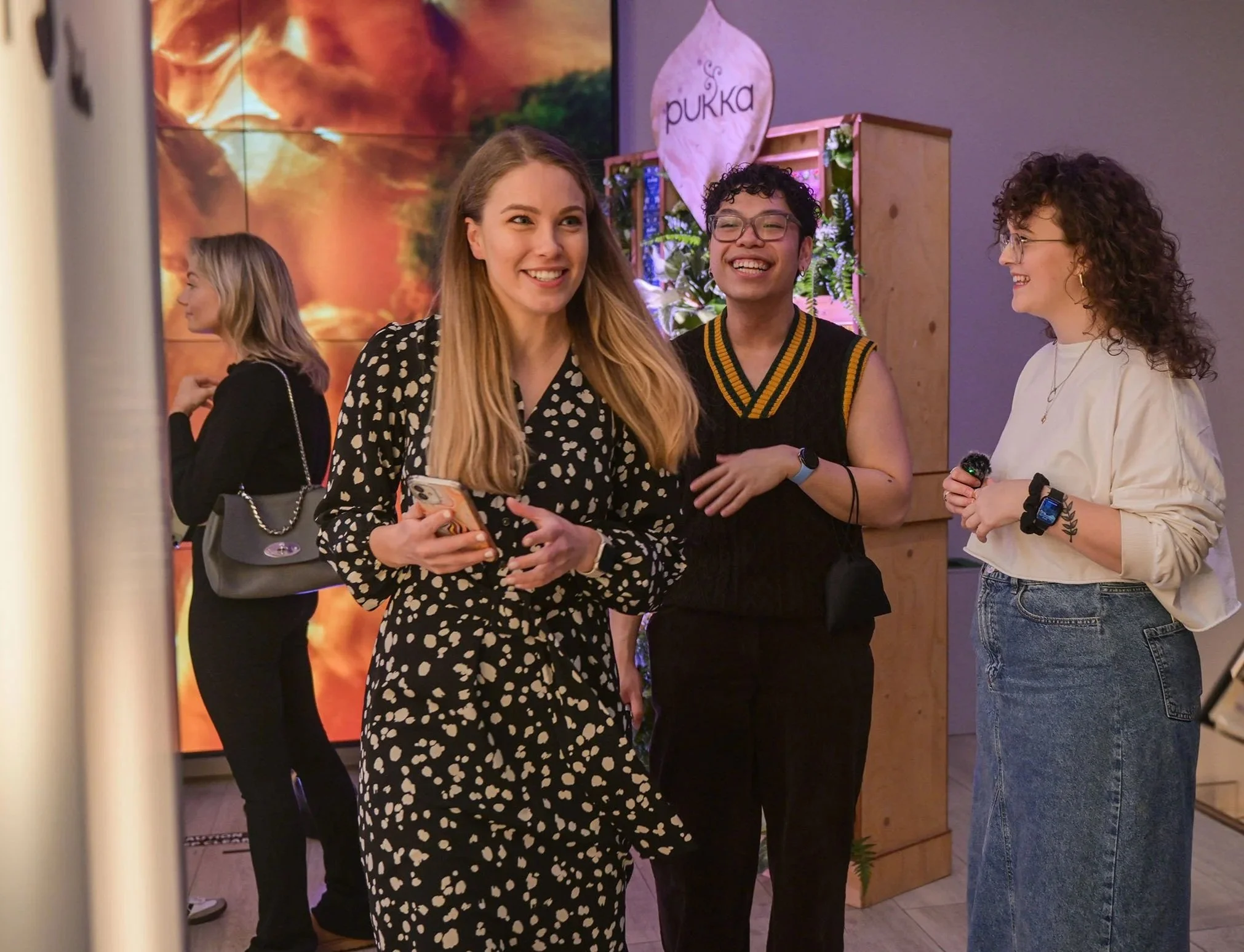 A group of three women smiling and talking at an indoor event. One woman is wearing a black dress with white polka dots, holding a phone. The second woman is in a sleeveless black top with a yellow stripe, and the third woman is in a white sweater and blue jeans. There is a woman in the background with blonde hair wearing black, and a decorative sign with the word "pukka" and a plant behind them.