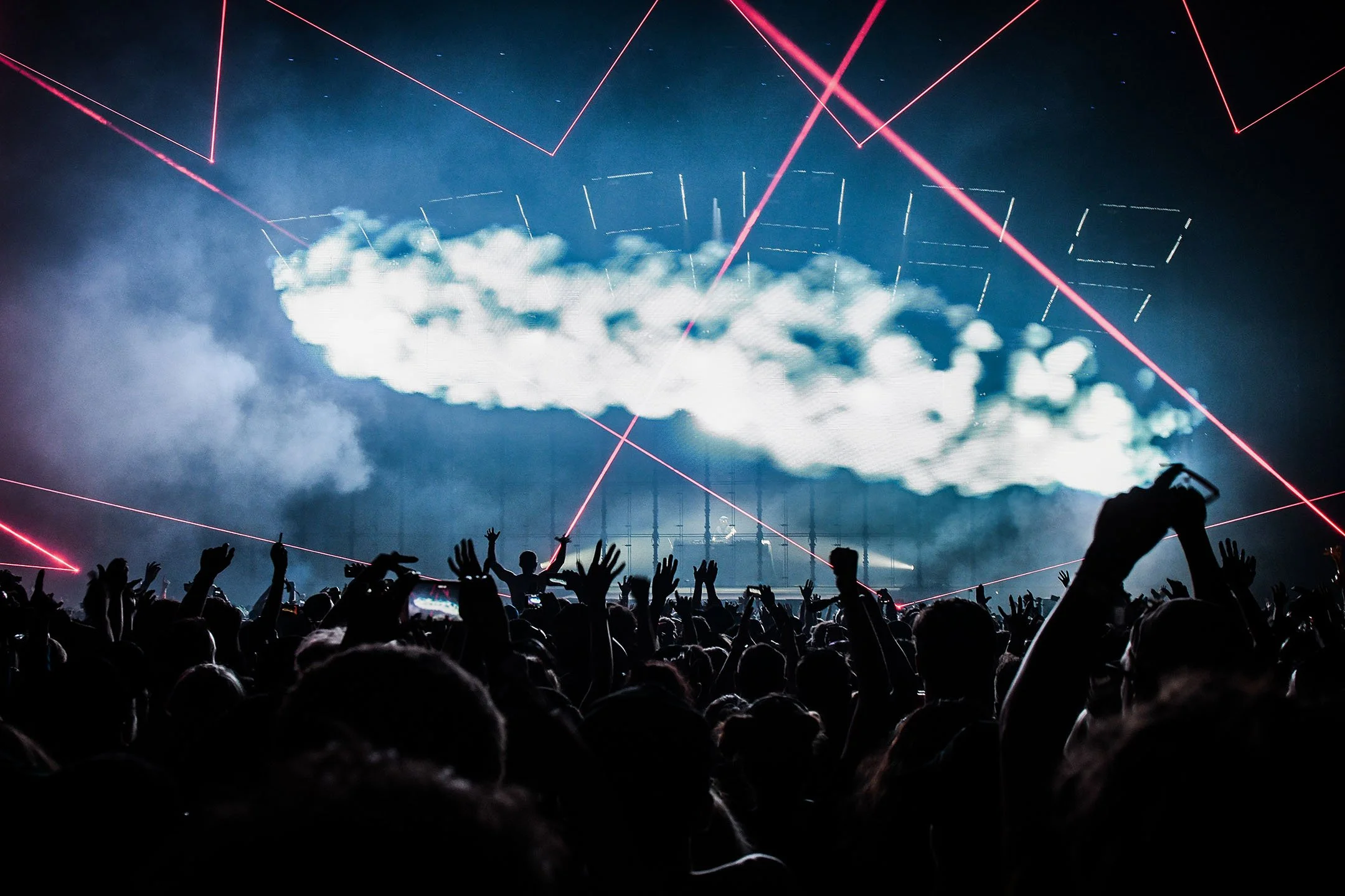 A concert with a crowd of people raising their hands, a stage with clouds, laser lights, and a DJ.
