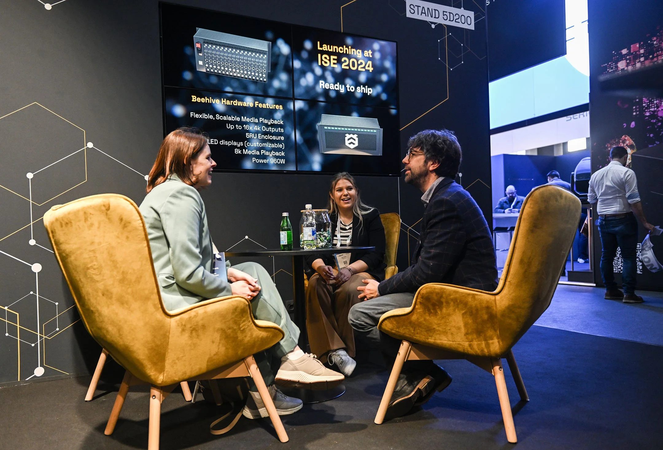 Three people sitting around a small round table, two women and one man, engaged in conversation at an event with a large digital screen behind them displaying information about a product launch for ISE 2024. The screen has text listing beehive hardware features and an image of hardware. Other attendees are visible in the background.
