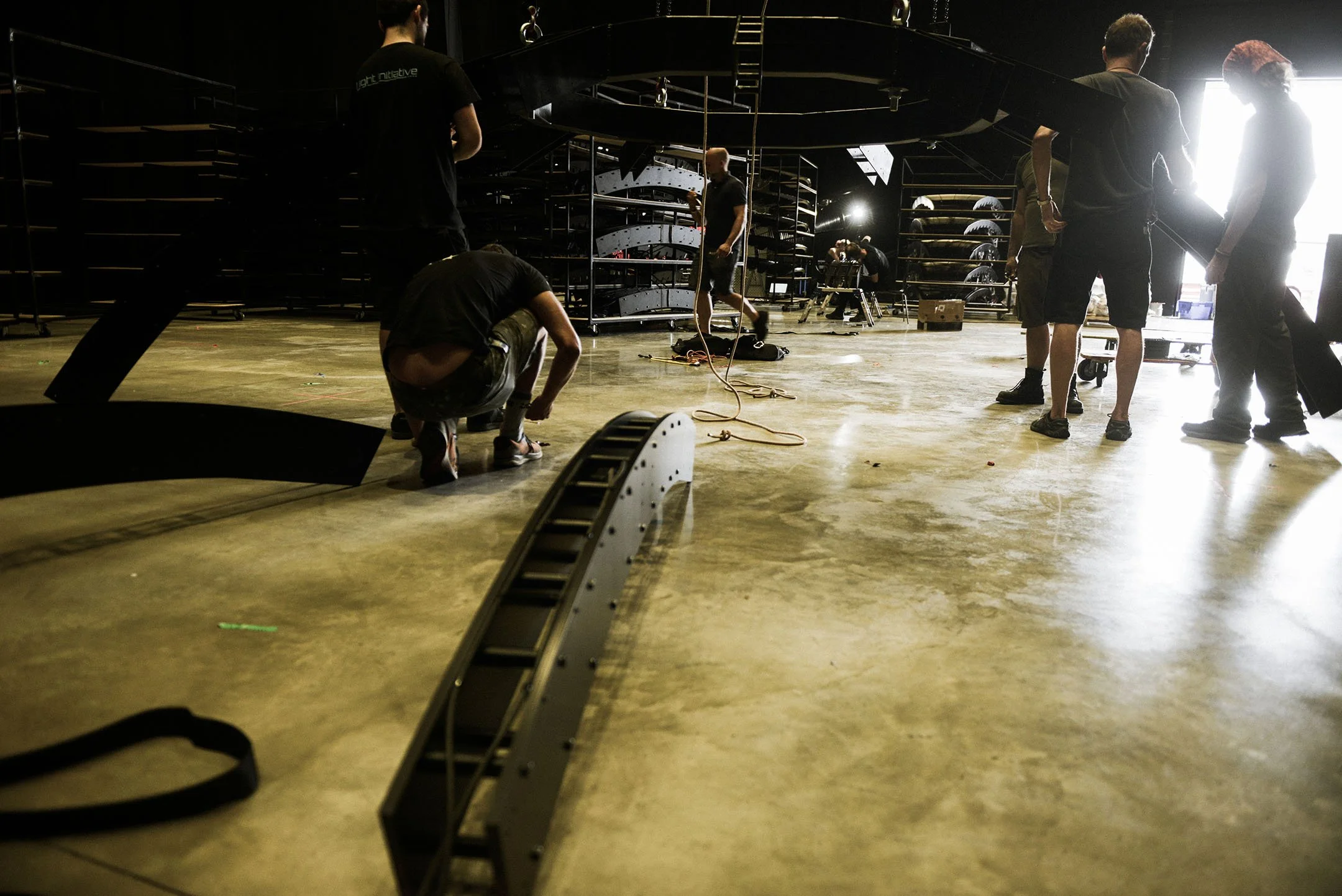Group of people assembling structures with black metal parts in a spacious industrial room, some crouching and some standing, with equipment and shelving in the background.