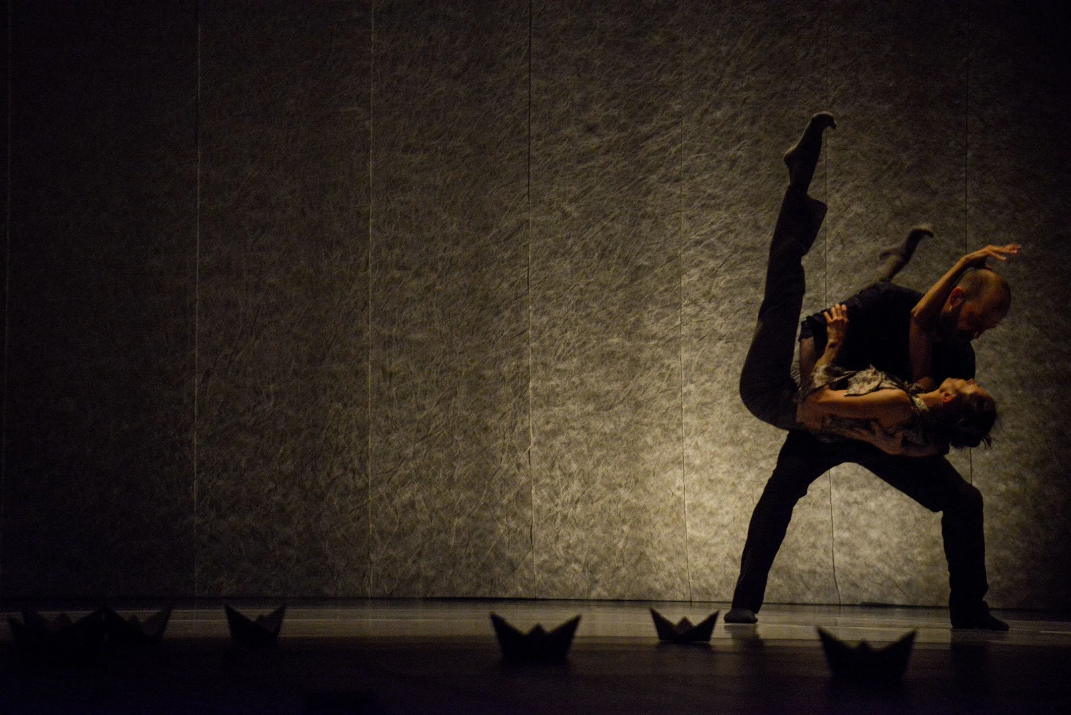 A dance performance featuring a pair of dancers, one supporting the other in an intricate pose, against a textured wall with low lighting.