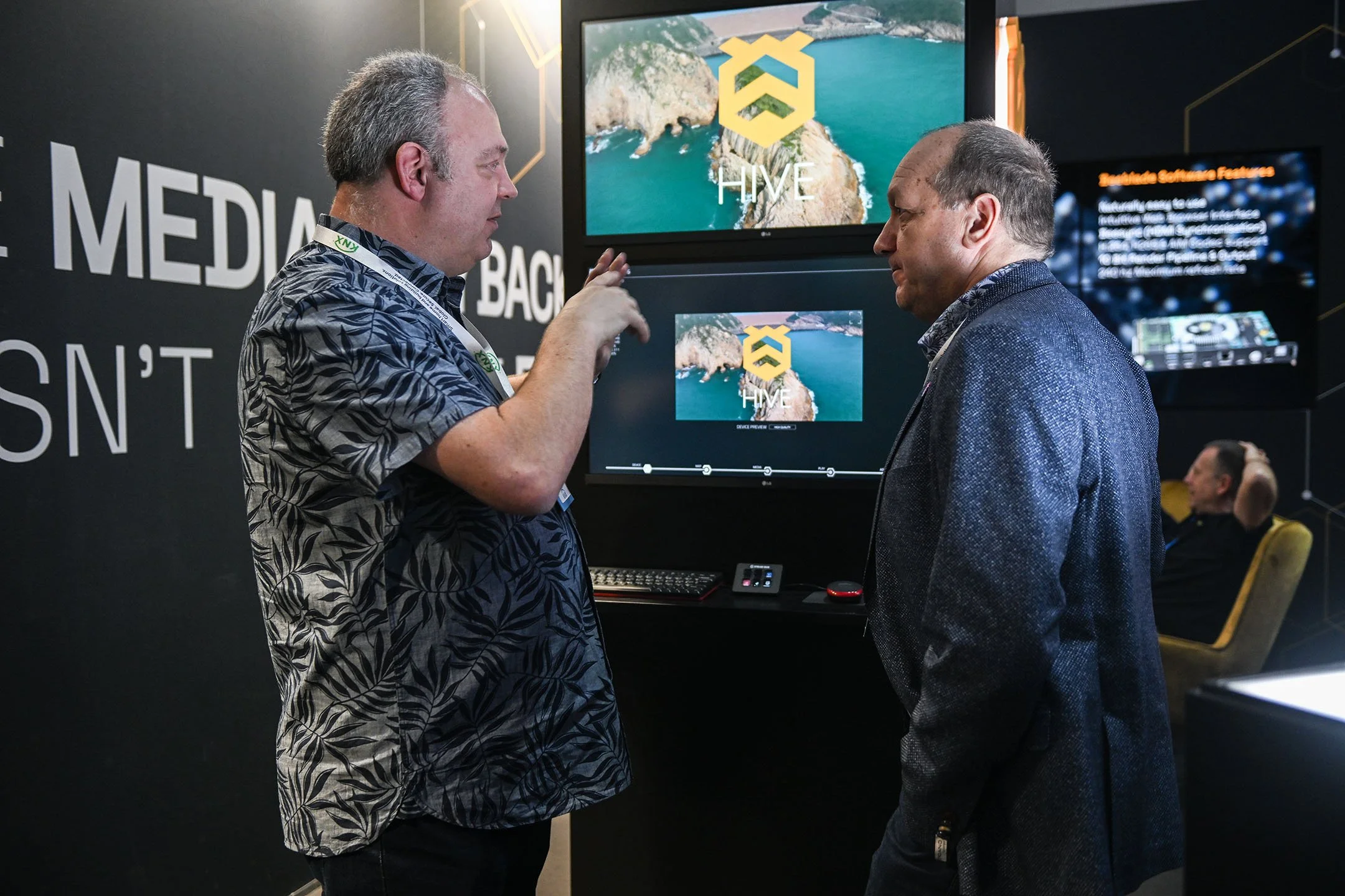 Two men are engaged in conversation at a technology exhibit. One man is gesturing with his hand while the other listens. Behind them are screens displaying the Hive logo and landscapes, along with a booth setup.