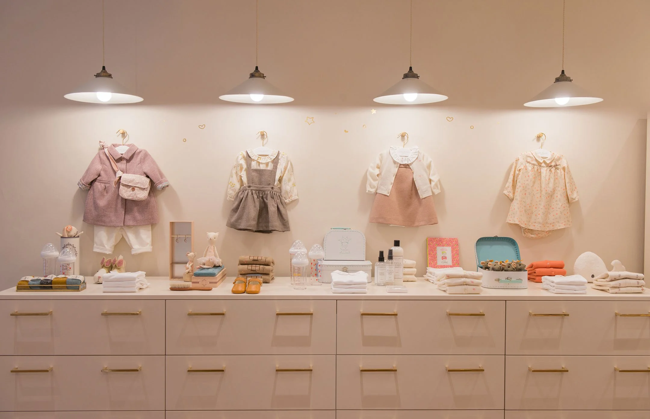 Display of children's clothing and accessories on a white dresser with four hanging outfits and various items such as toys, folded towels, and bottles.