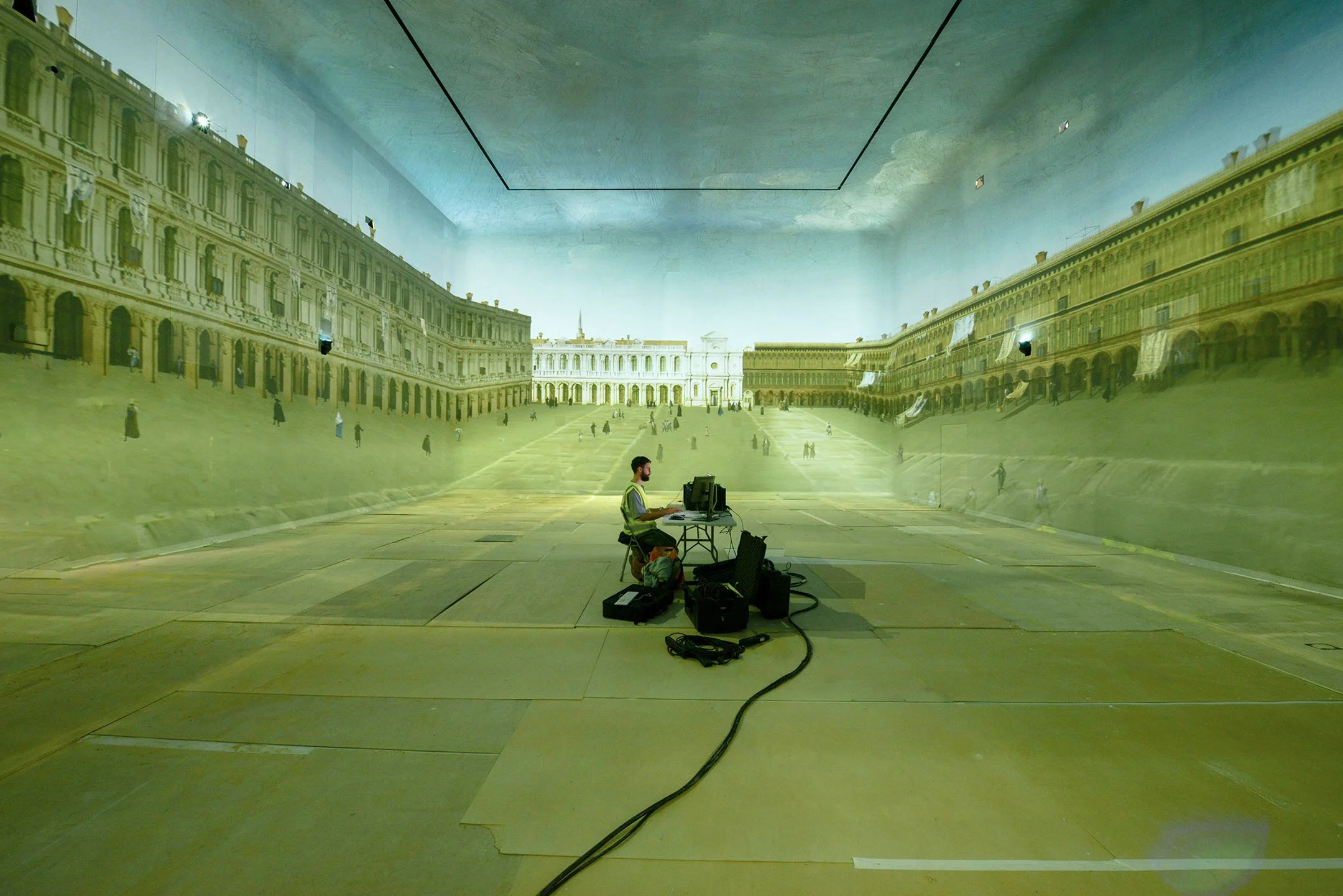 A person working on a computer in a large room with a projected image of a historic building and courtyard covering the walls and floor.