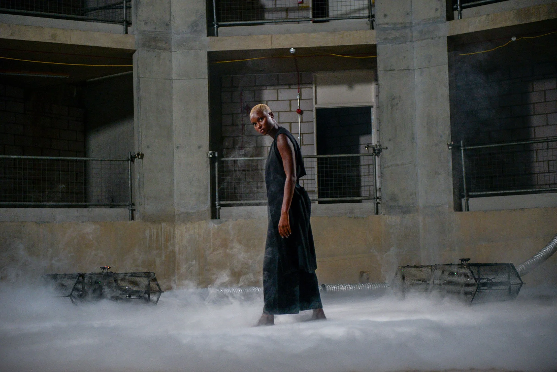 A woman with short blonde hair and dark skin stands barefoot in a foggy industrial-looking space, wearing a black sleeveless dress and looking over her shoulder.