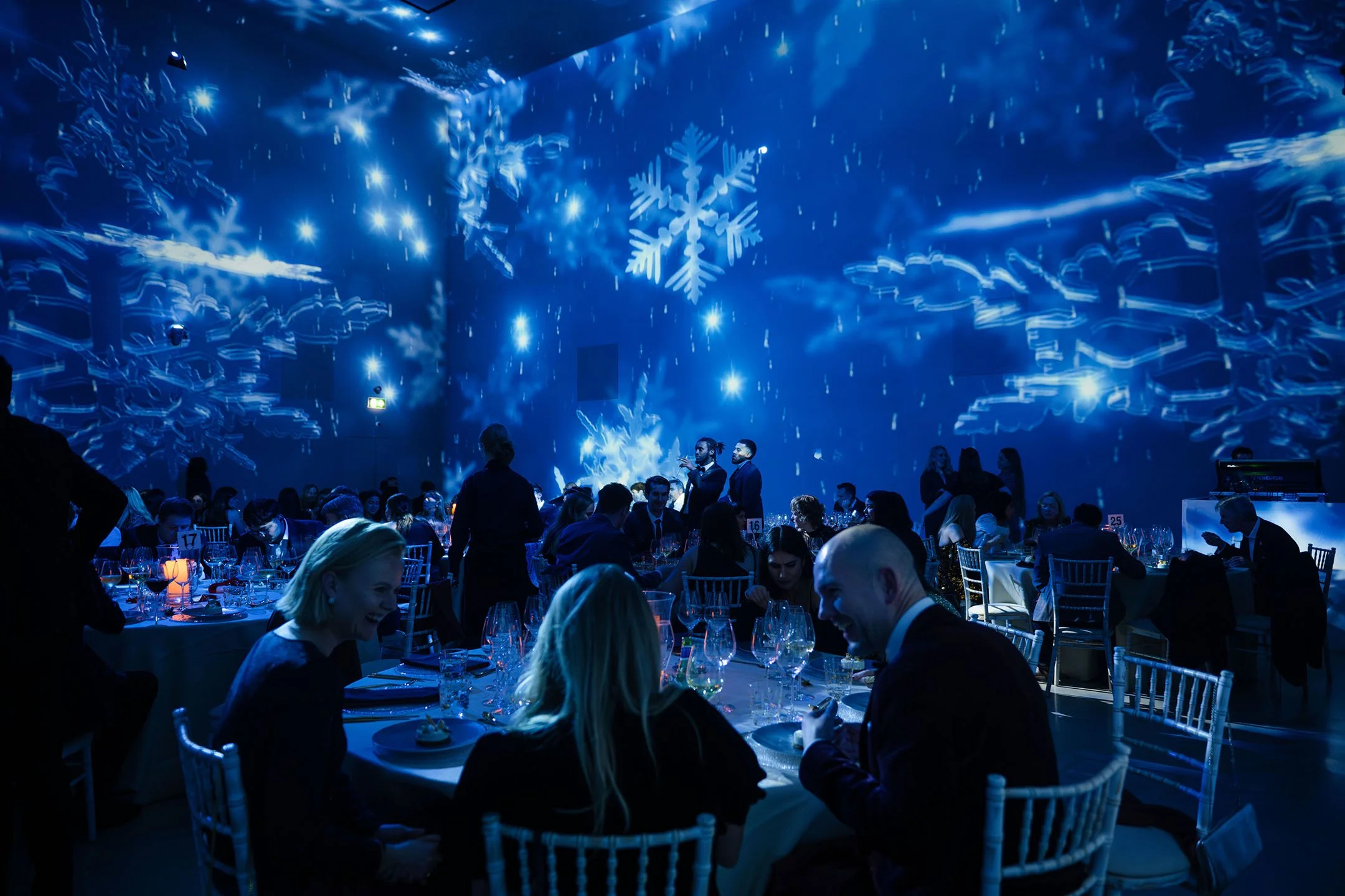 Guests seated at round tables in a dimly lit room with blue lighting and projected snowflake decorations, celebrating at an event.