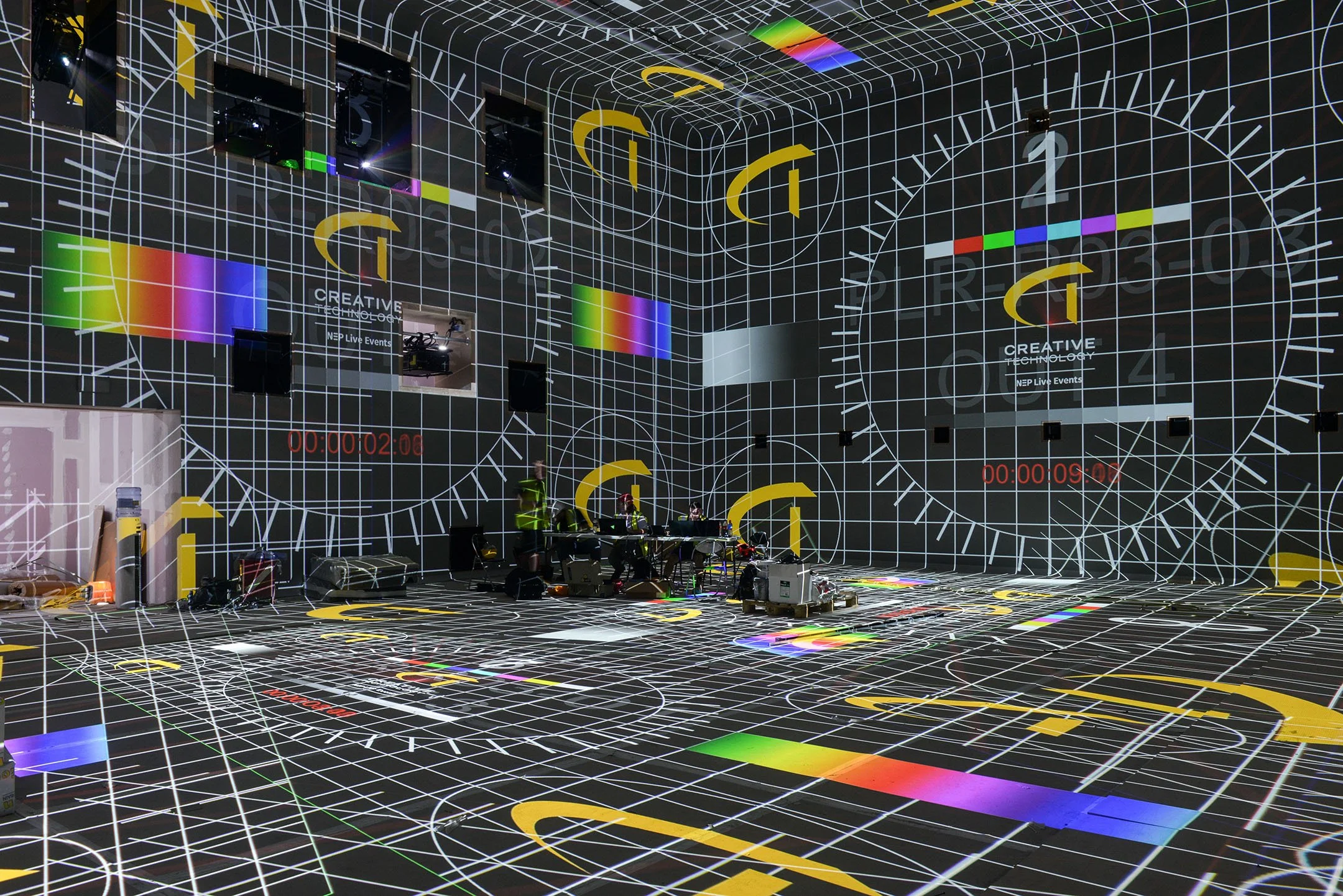 grid pattern projected on walls and floor, featuring geometric lines, circles, and colorful bars, creating a digital or futuristic environment, with some equipment and individuals sitting at a table in the center. photo © Antonio Pagano