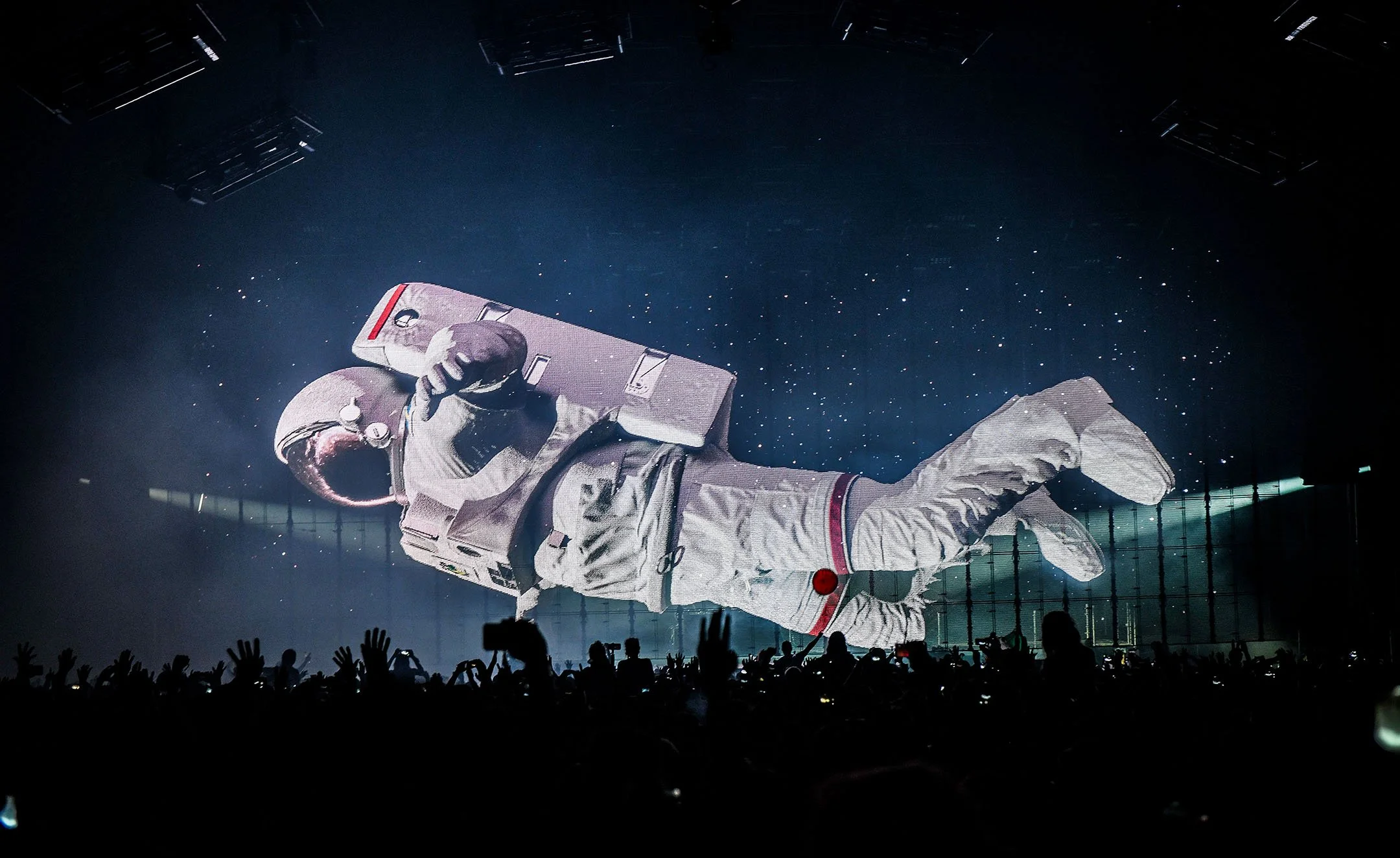 A giant screen displaying an astronaut in a space suit floating with one hand reaching out at a concert or event, with a crowd of people in front capturing the moment.