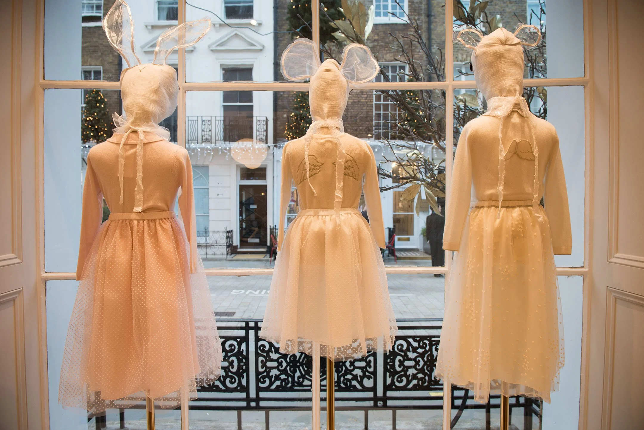 Three mannequins dressed in elegant cream-colored dresses with sheer overlays, arranged in a row behind a large window display. Each mannequin has a fabric head wrapped in matching cloth and wearing bunny or angel-themed headbands with sheer ears or wings. The background outside shows a street scene with decorated Christmas trees and buildings.