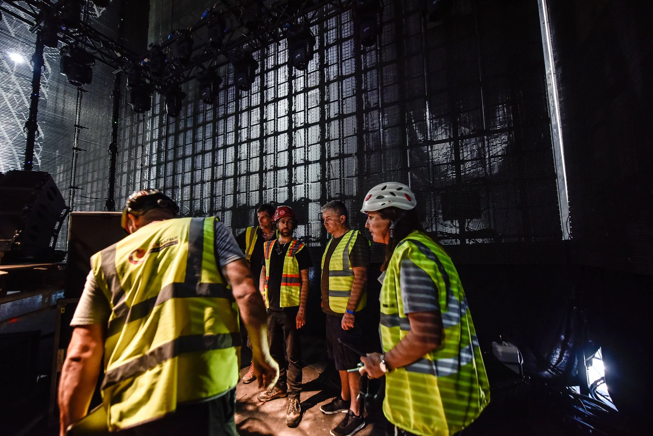 Group of workers in high visibility safety vests and helmets backstage at a concert or event.