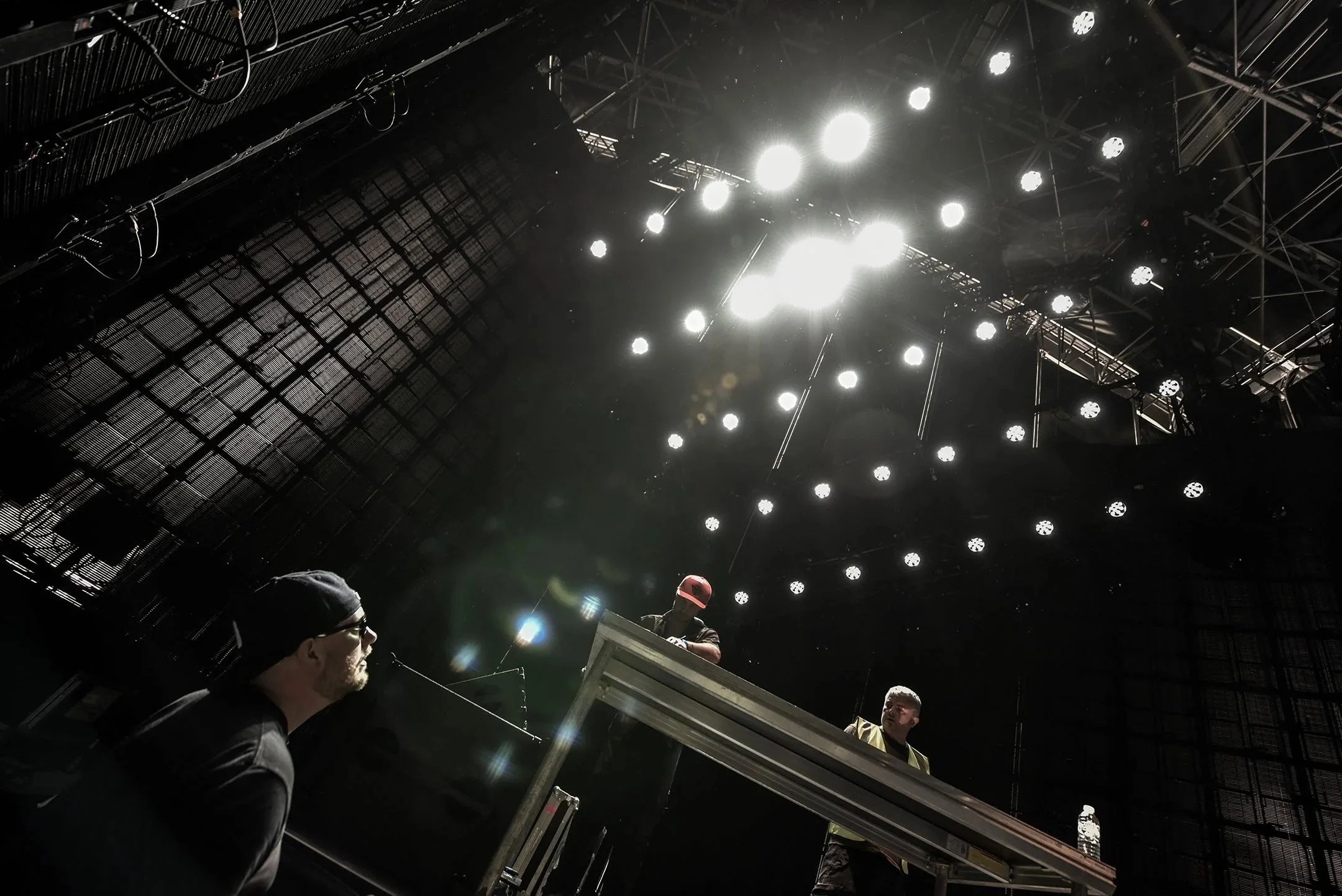 Two people at a stage or concert setup, with one standing behind a large equipment or table and the other sitting or leaning nearby, under bright stage lighting and a dark industrial-looking roof structure.