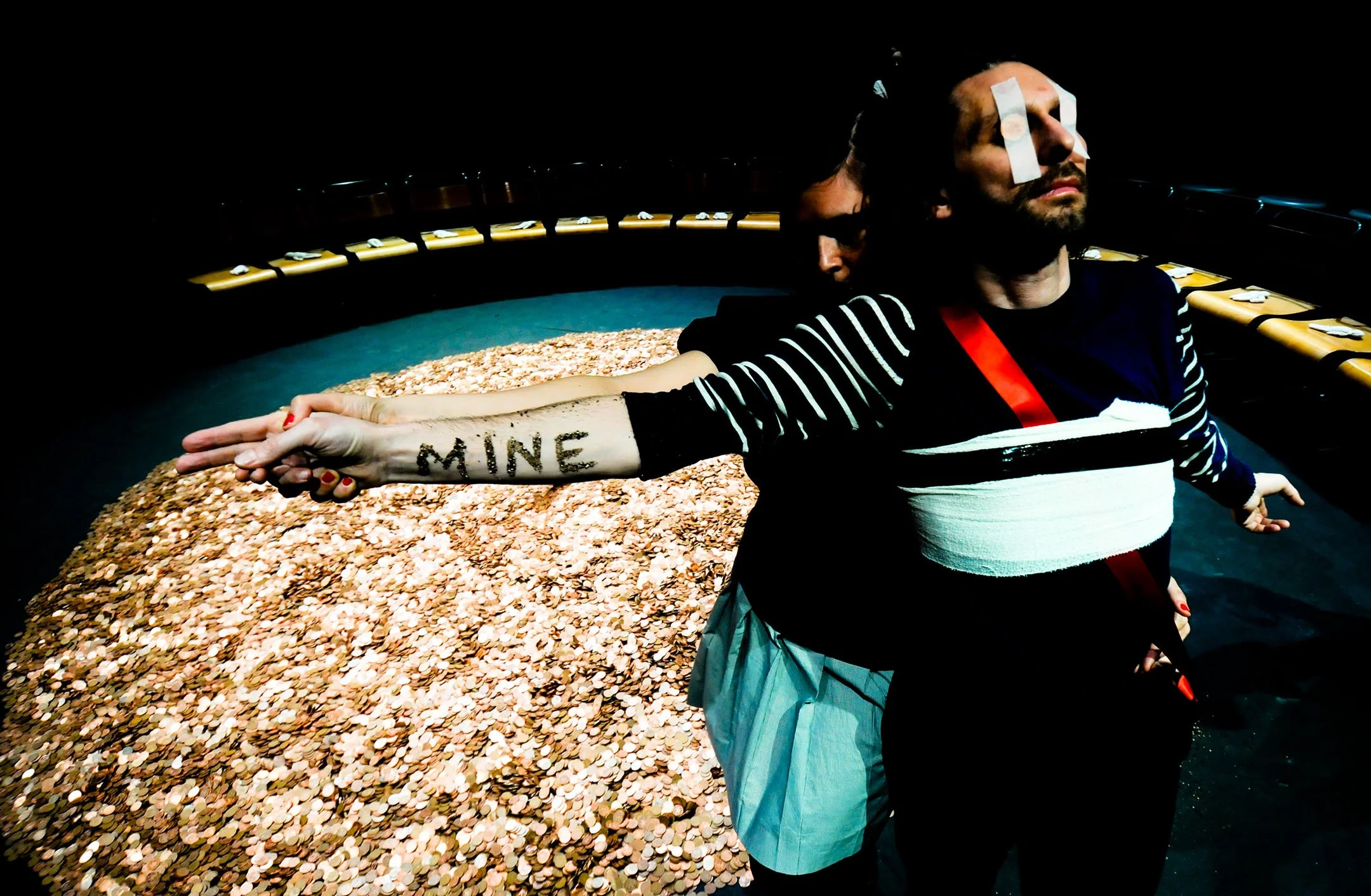 Two people with their arms linked, one with the word "MINE" written on their forearm, standing in front of a large pile of coins on a circular stage.