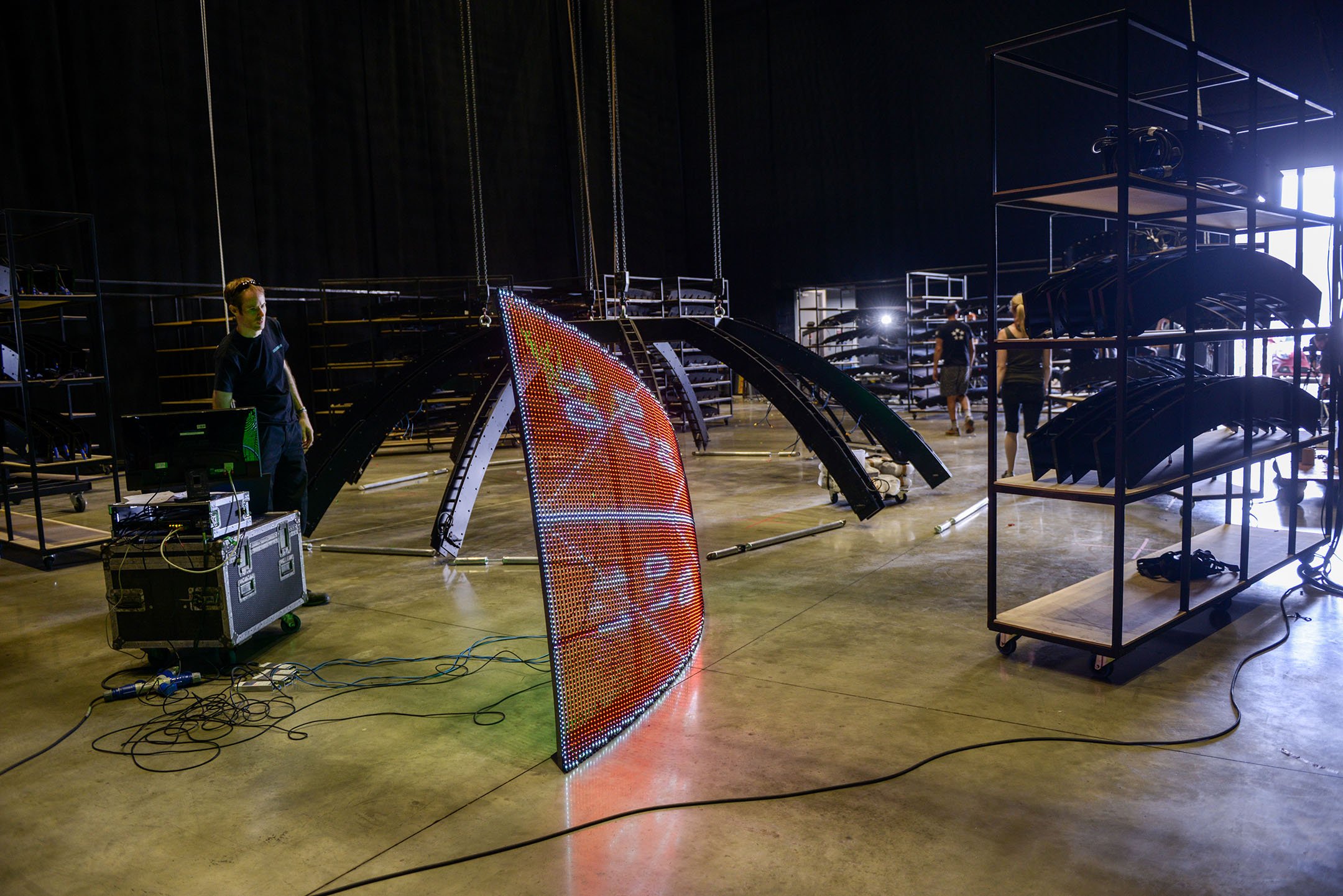 Interior view of a warehouse or event space with LED screens, metal racks holding equipment, and people setting up for an event.