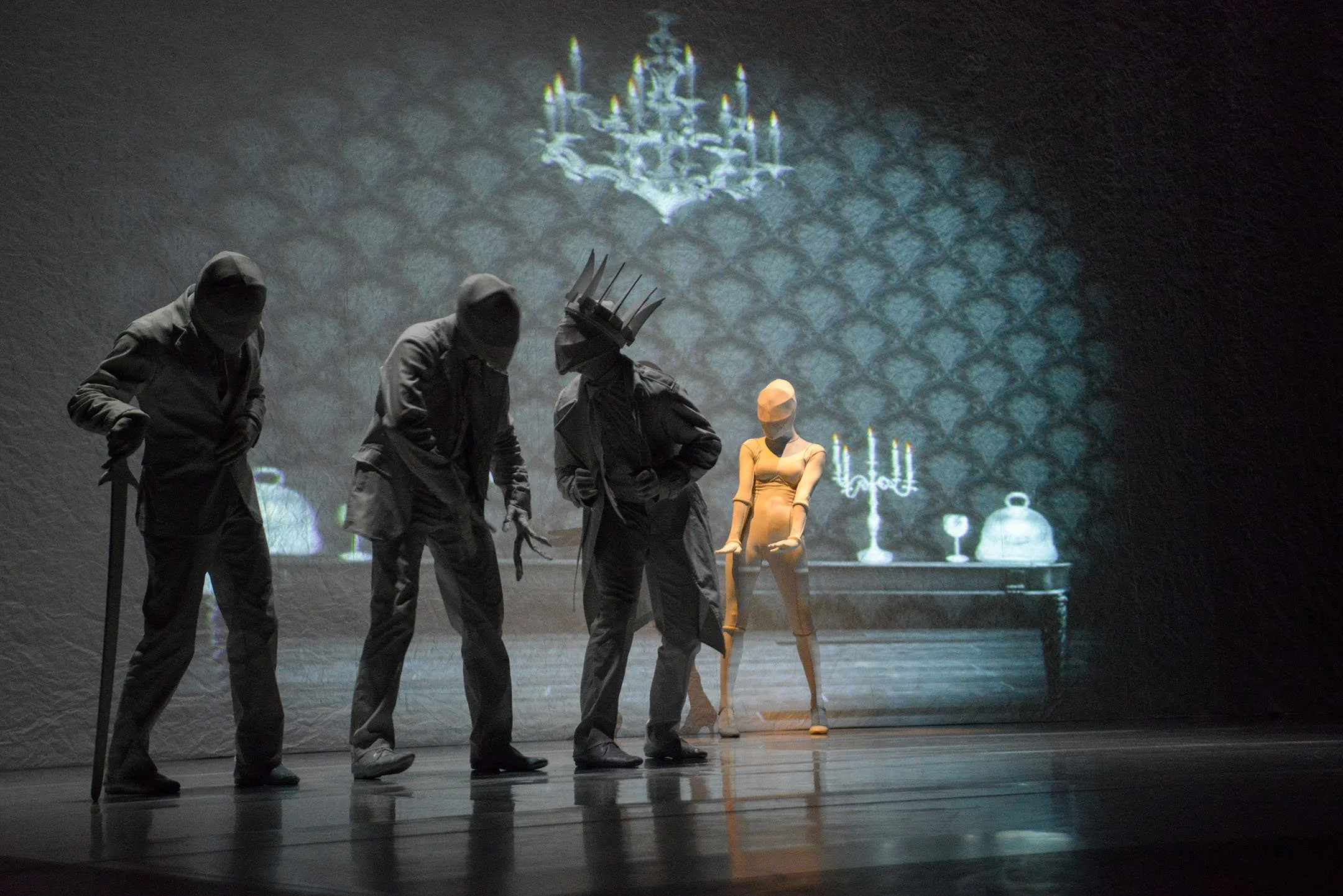 Four performers dressed in dark costumes and masks on stage, with a woman mannequins and a projected background of a chandelier and a decorative wall.