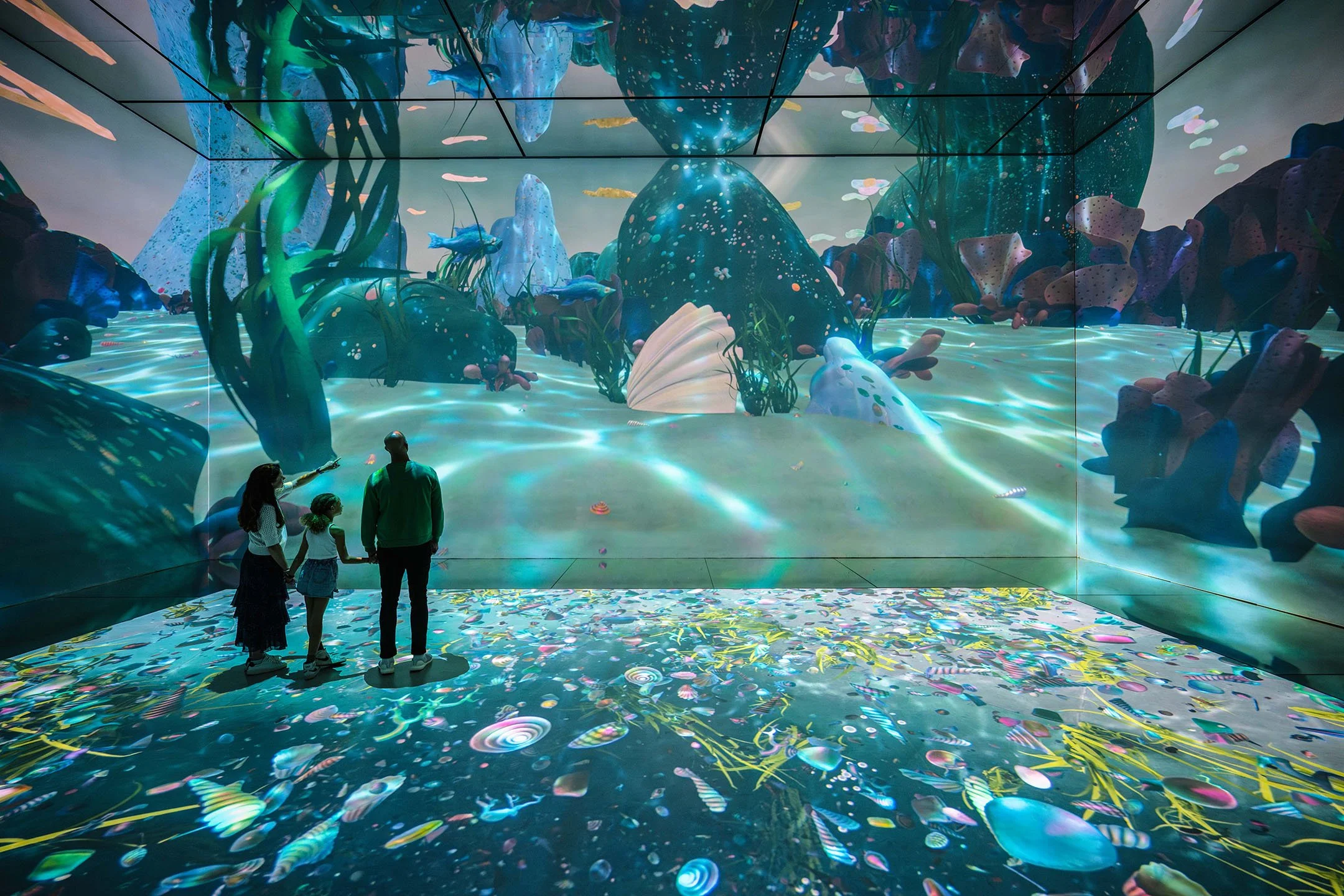 Family of four, including two children, standing in front of a large digital aquarium display with colorful coral and fish.