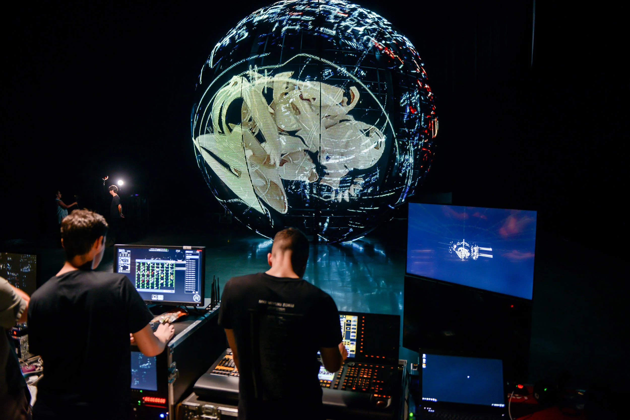 Sound engineers working on sound equipment in front of a large digital globe projection displaying an animated human skeleton, in a dark performing arts space.