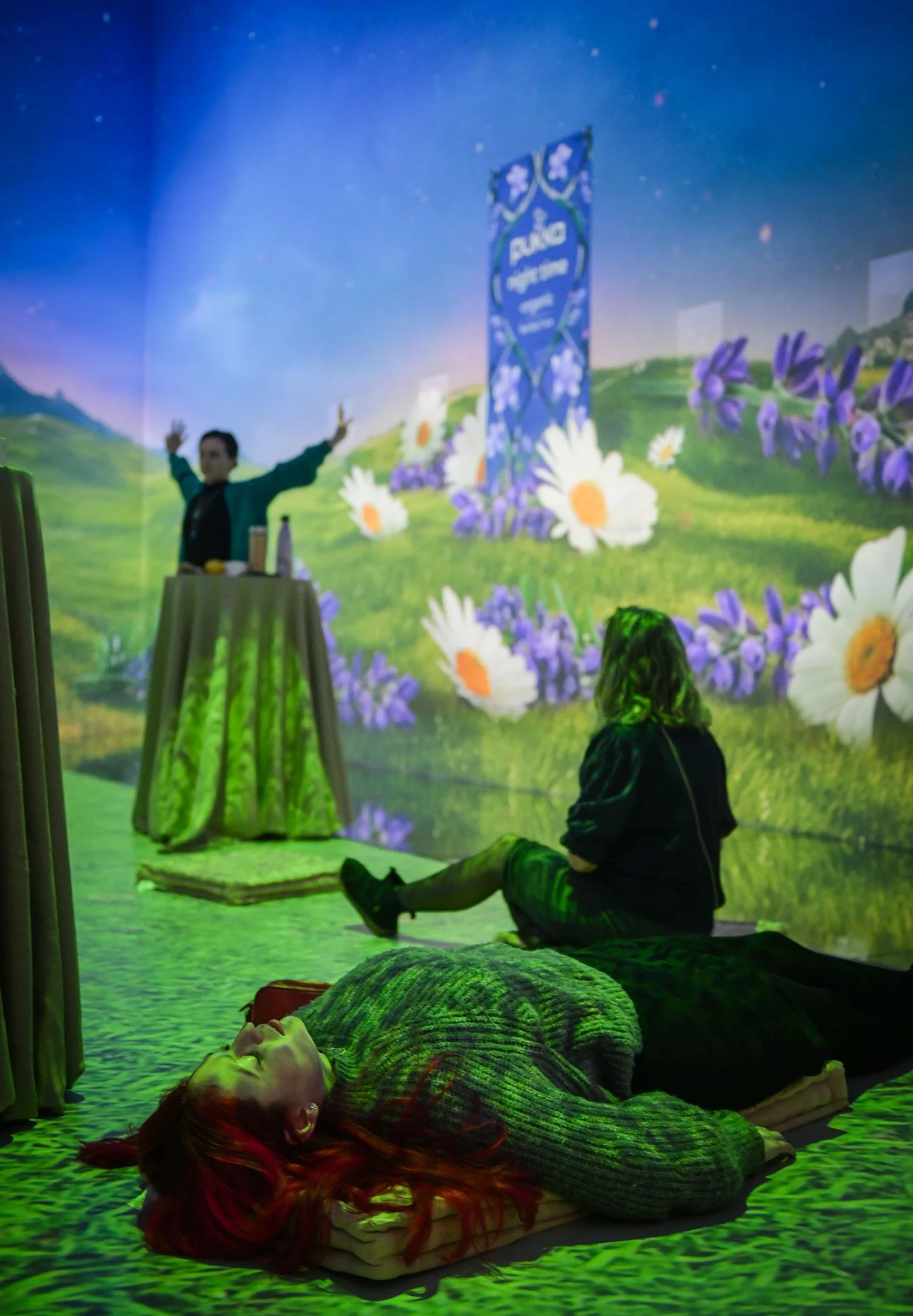 An indoor scene with two women lying on the floor, one with red hair resting on a pillow and the other with hairstyle obscured sitting up, observing a person performing on a stage with a colorful, floral, animated backdrop.