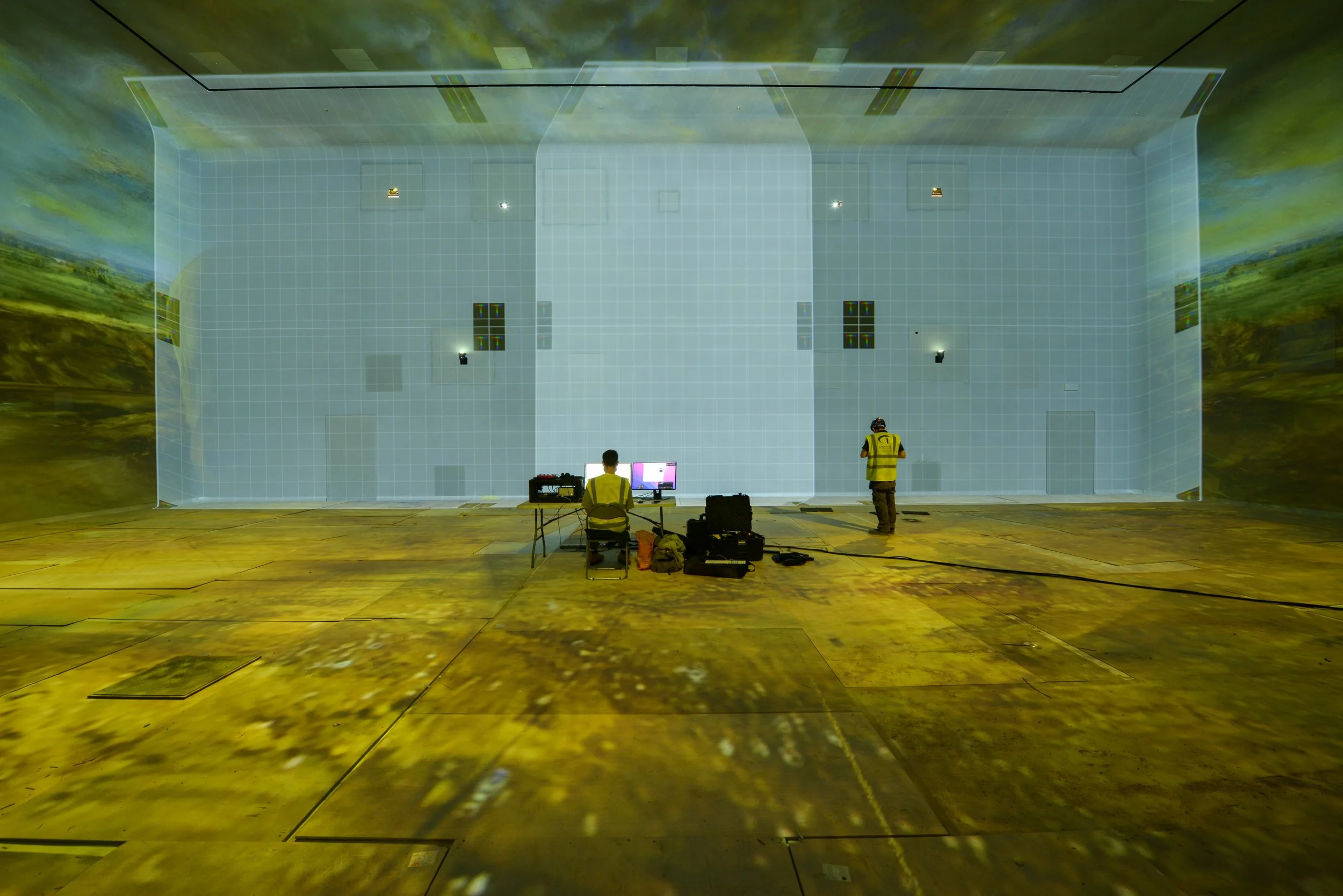 Two individuals working in a large immersive room with digital projection on the walls and floor, with equipment and computers set up in front of them. The making of Frameless museum photo © Antonio Pagano