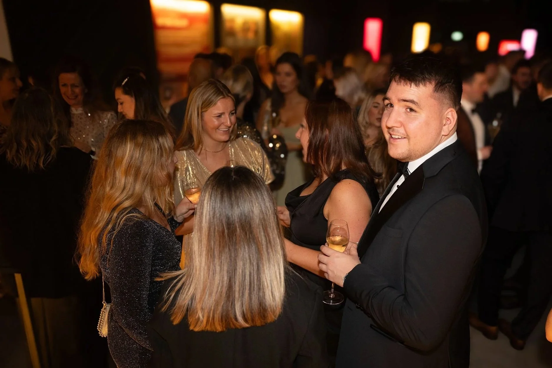 Group of people dressed in formal attire at a social event, holding glasses of wine, in a dimly lit setting with colorful lights in the background.