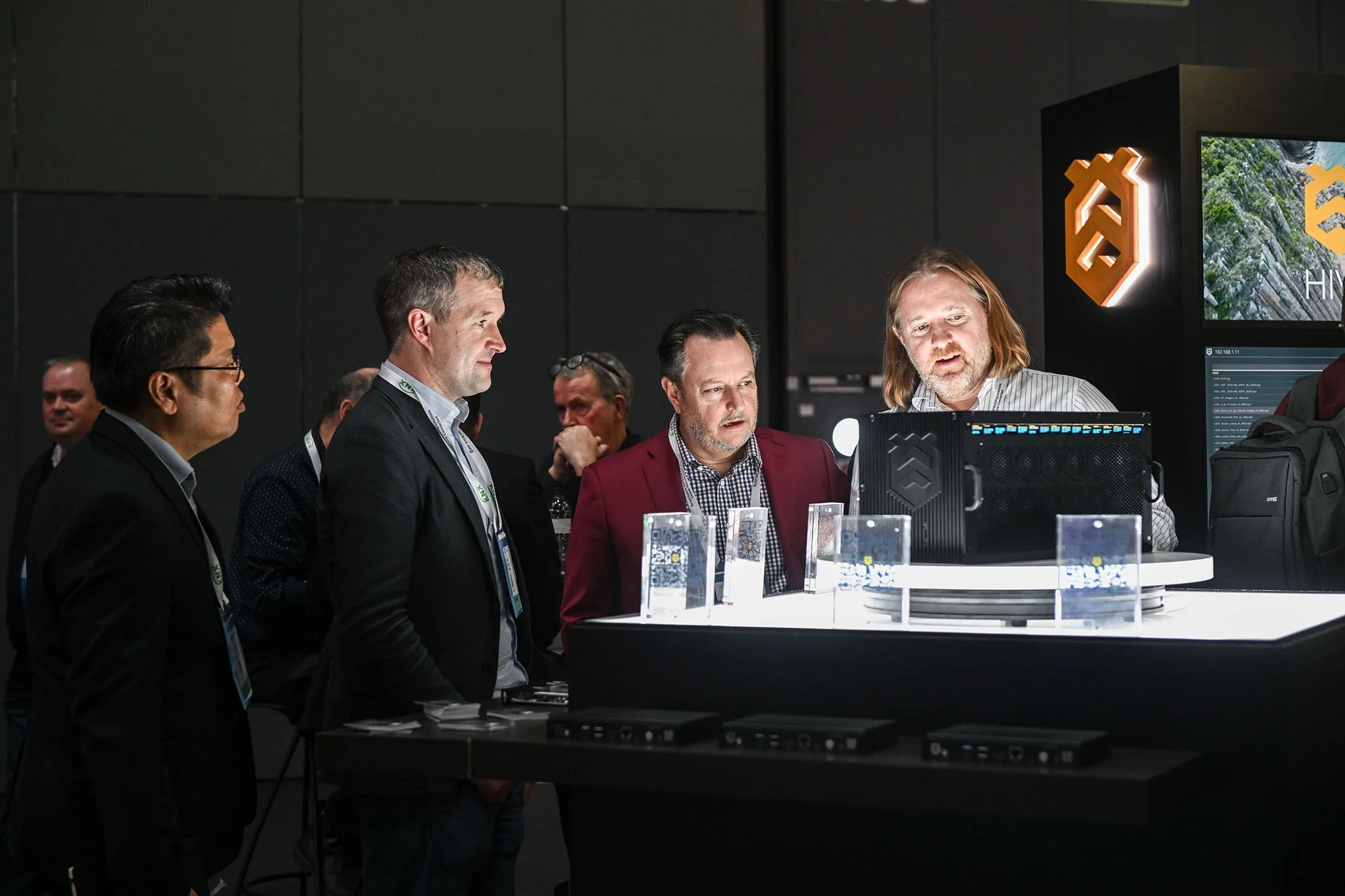 Group of men gathered around a display at a tech conference, observing a computer, with a dark background and illuminated signage.