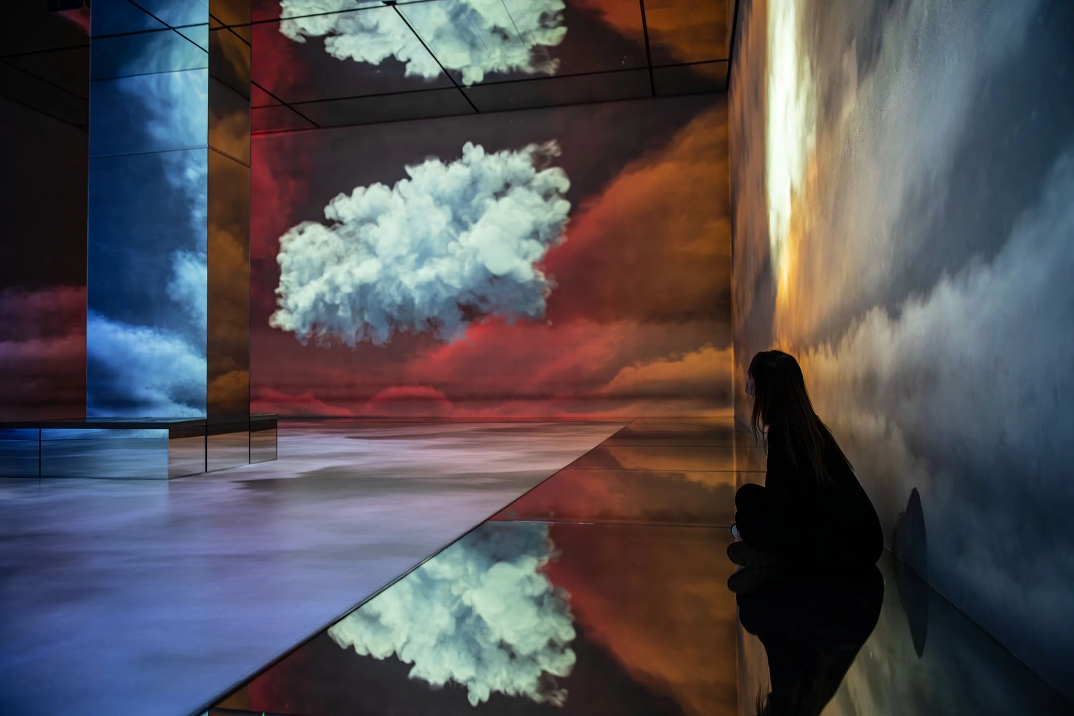 A woman sitting on the floor in an art gallery with large projected images of clouds and sky on the walls and floor, creating a reflection effect.