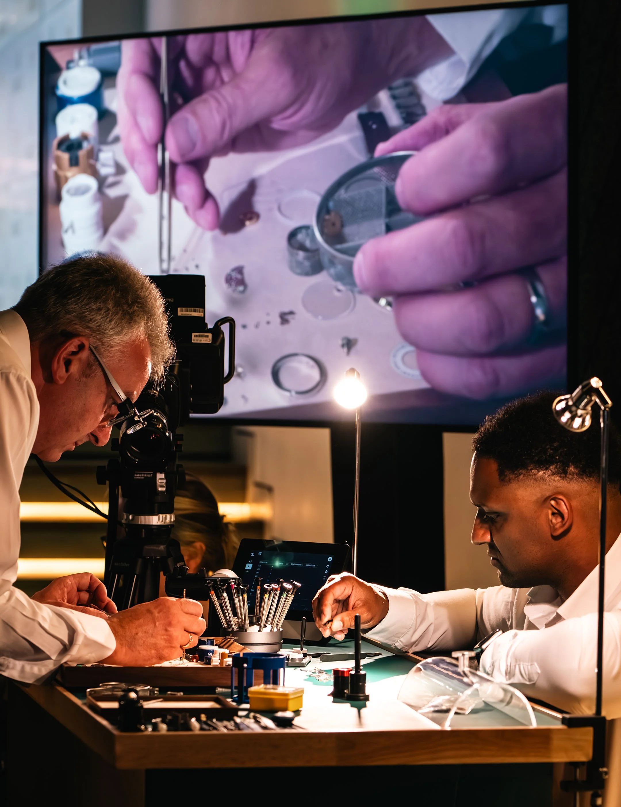 Rolex launch & gala dinner by DMR (David M Robinson-Jewellery & Watches) at Frameless immersive museum, London. Two watchmakers working disassembling a Rolex watch , with a large screen above showing a close-up of watch parts.