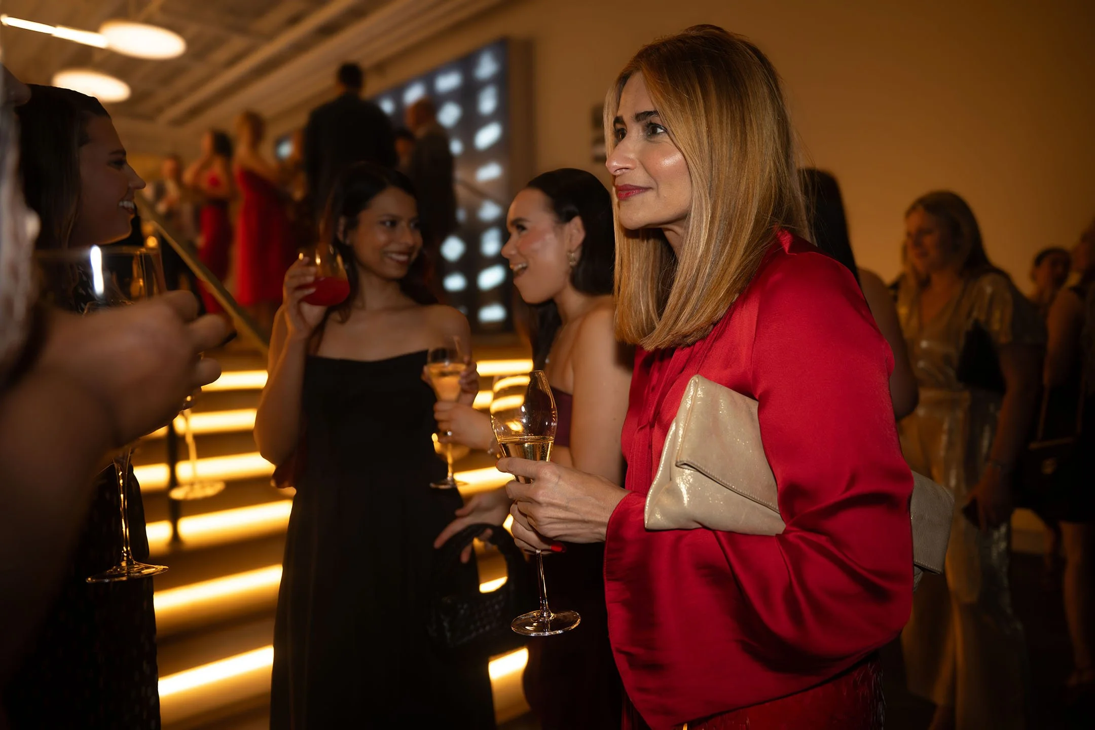 Group of women at a social event, holding glasses of champagne, engaging in conversation, with some smiling and others listening; warm lighting and modern interior decor.