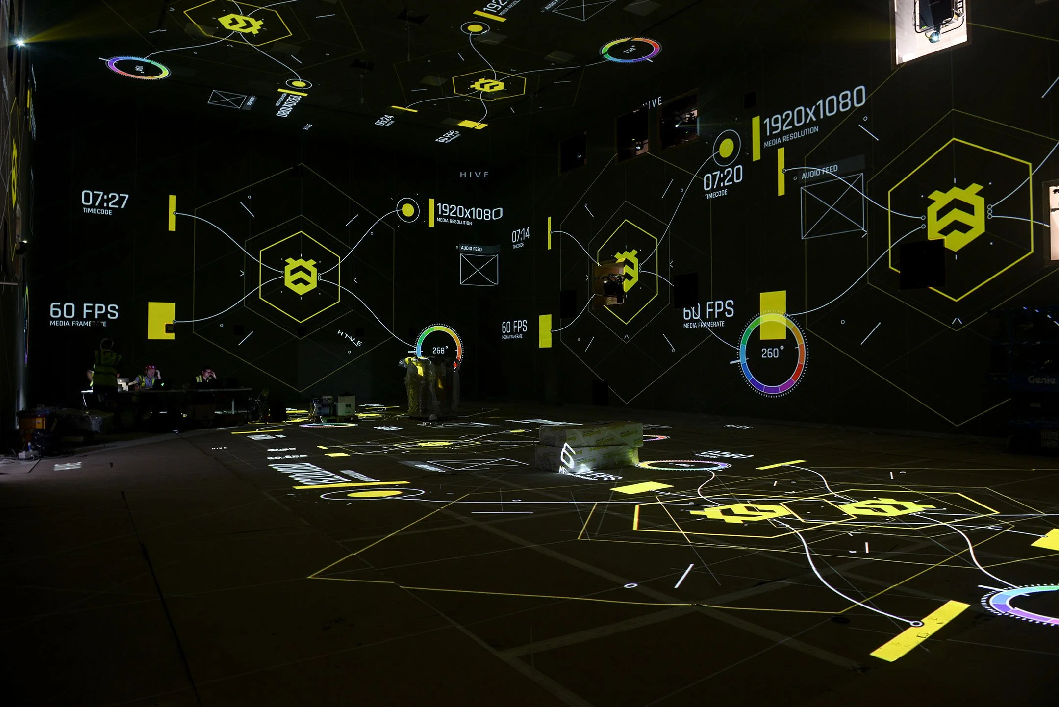A dark room with digital graphic projections on the walls and floor, displaying technical data, graphs, and symbols in yellow, white, and other colors, resembling a futuristic interface or a media production studio.  photo © Antonio Pagano