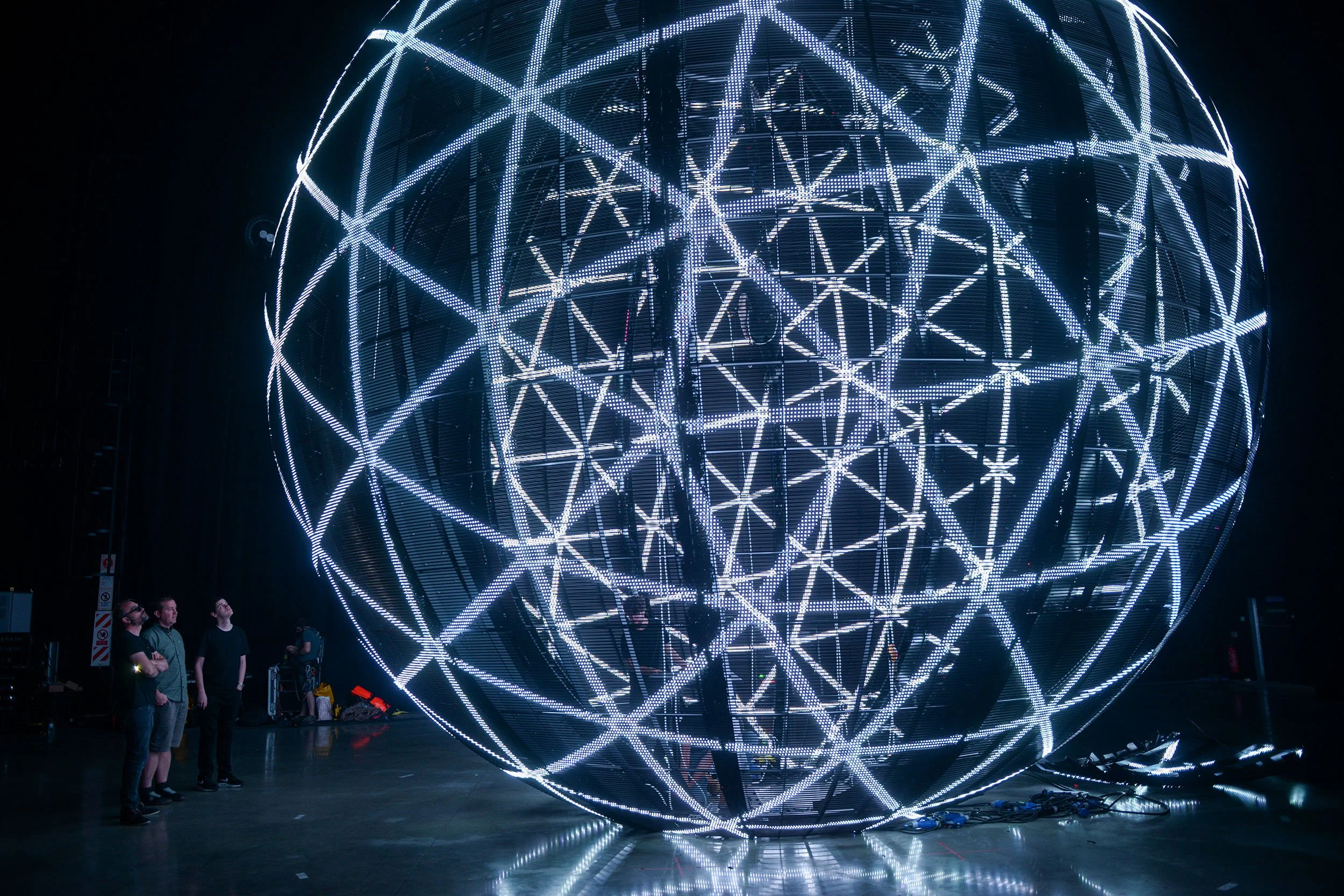 A large spherical LED light installation with geometric patterns, illuminated in white and blue, in a dark indoor space. Several people are standing nearby, observing the structure.