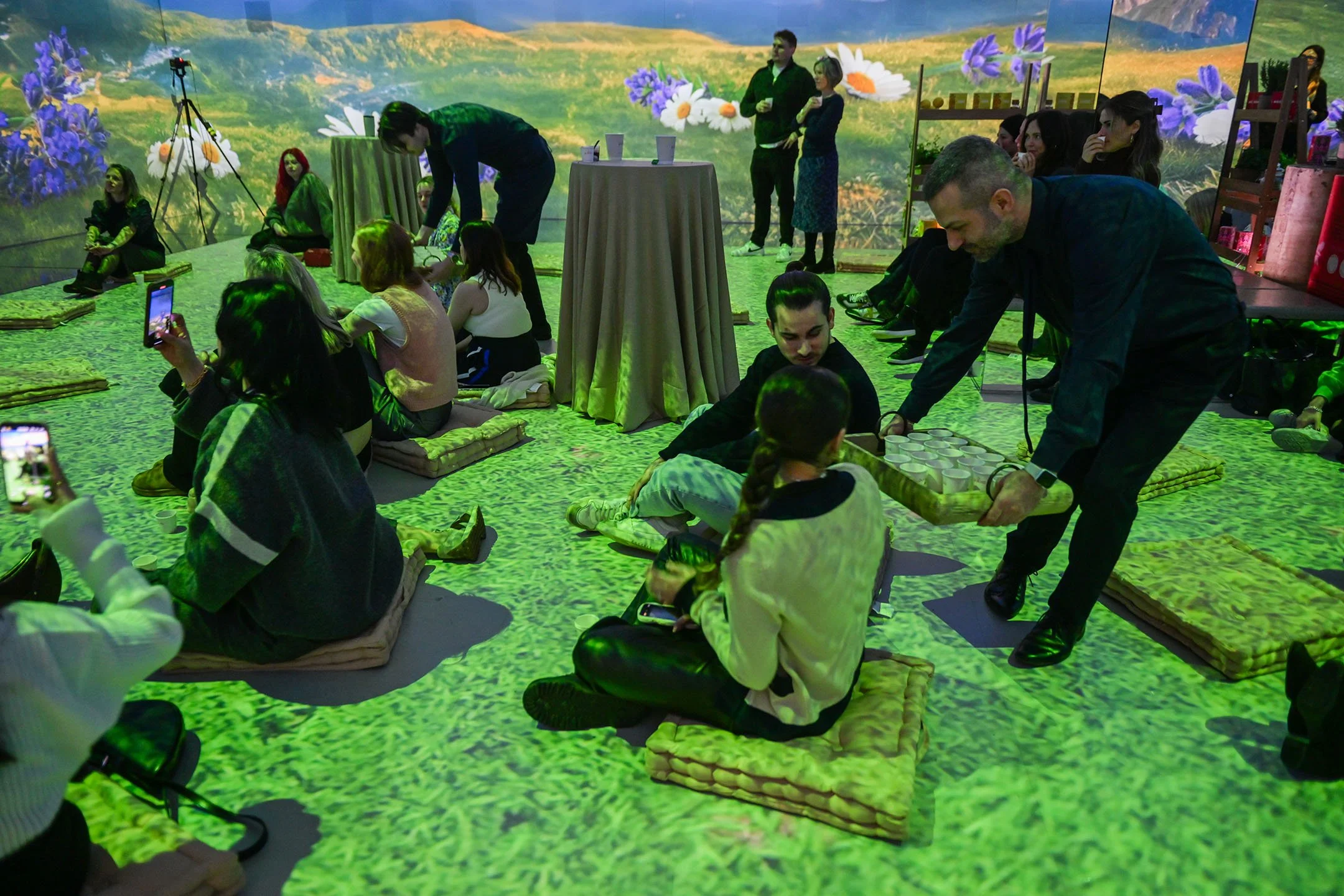 People sitting on cushions on the floor, some taking photos, in a room with a digital landscape projection of a field of flowers on the walls, and a man serving drinks to a woman.