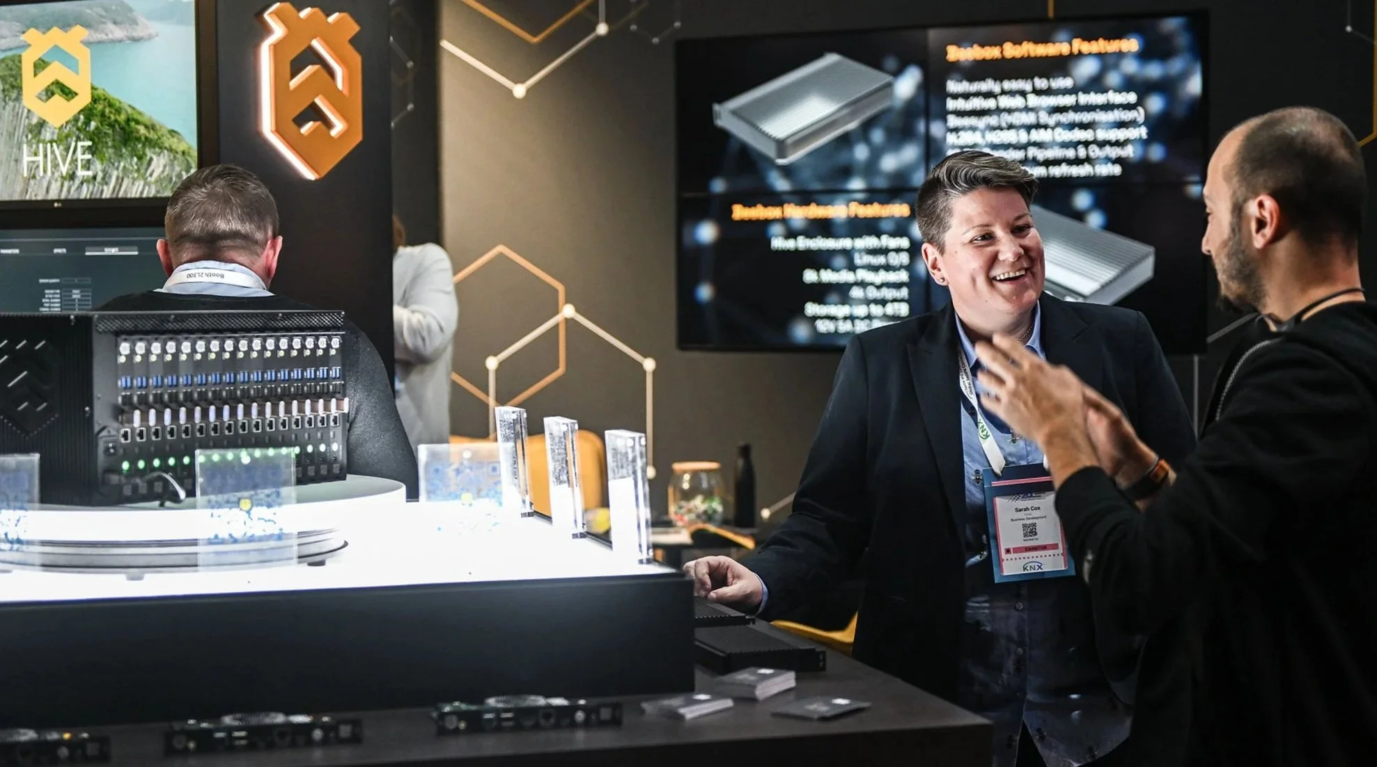 Two women are talking at a technology exhibit with other people and displays in the background. One woman is smiling and wearing a badge, and there is a large screen behind them showing product information.