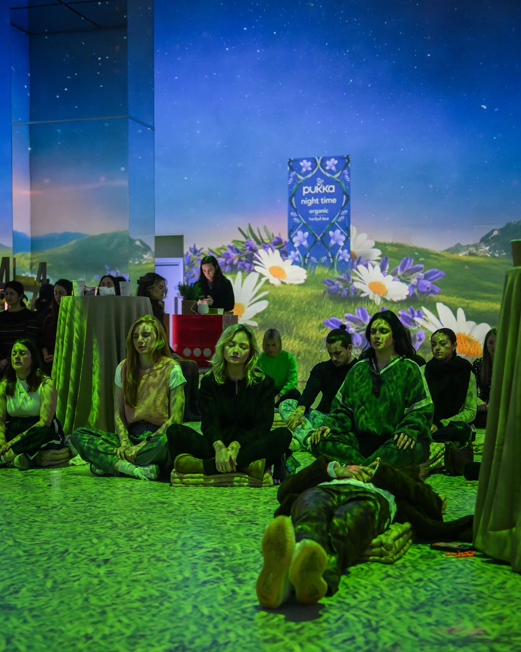 Group of women sitting cross-legged on the floor during meditation or prayer session, with a person lying down in front of them, illuminated by green lighting, in a room with a large digital projection of flowers and a blue sky background.