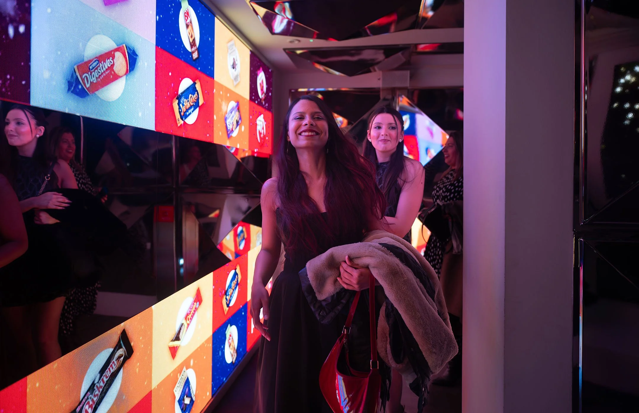 A group of smiling women at an event, standing in front of a colorful digital display with snack food advertisements, including Oreo, Chips Ahoy!, and Reese's, in a modern setting with reflective surfaces and vibrant lighting.