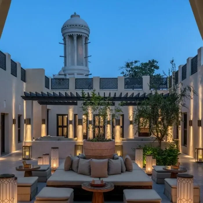 Chedi outdoor courtyard  in Sharjah at dusk with modern furniture, large cushions, and decorative lanterns, surrounded by a white stucco wall and greenery, with a tall tower and blue sky in the background.