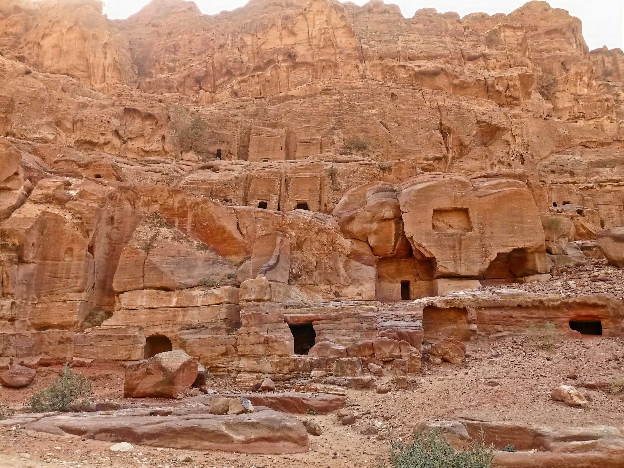 Ancient rock-cut tombs carved into a reddish-brown cliff face in Petra, Jordan, with multiple openings and narrow pathways.