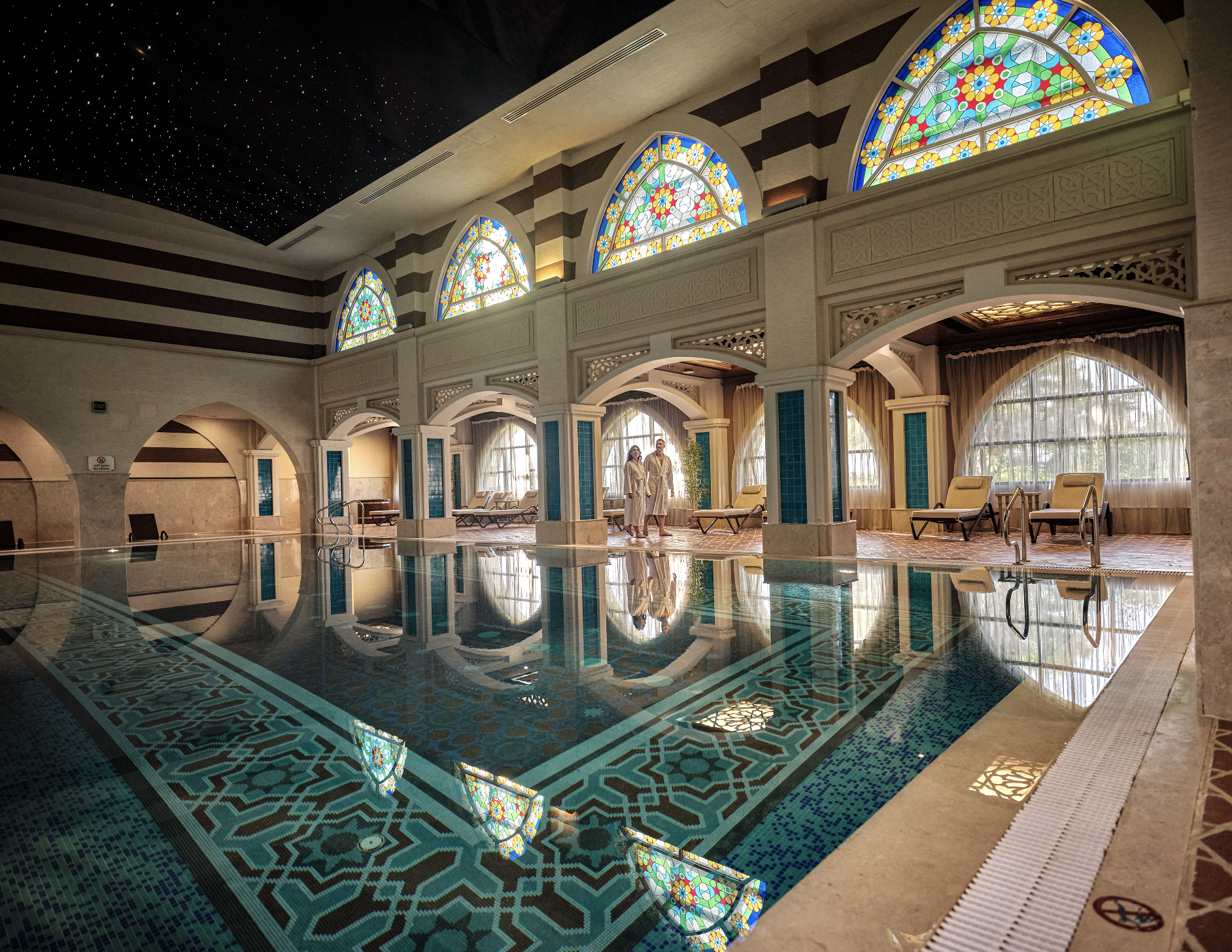 Interior of a luxurious indoor swimming pool area with ornate stained glass windows, a starry ceiling, and relaxed lounge chairs, with two women in bathrobes walking along the poolside.