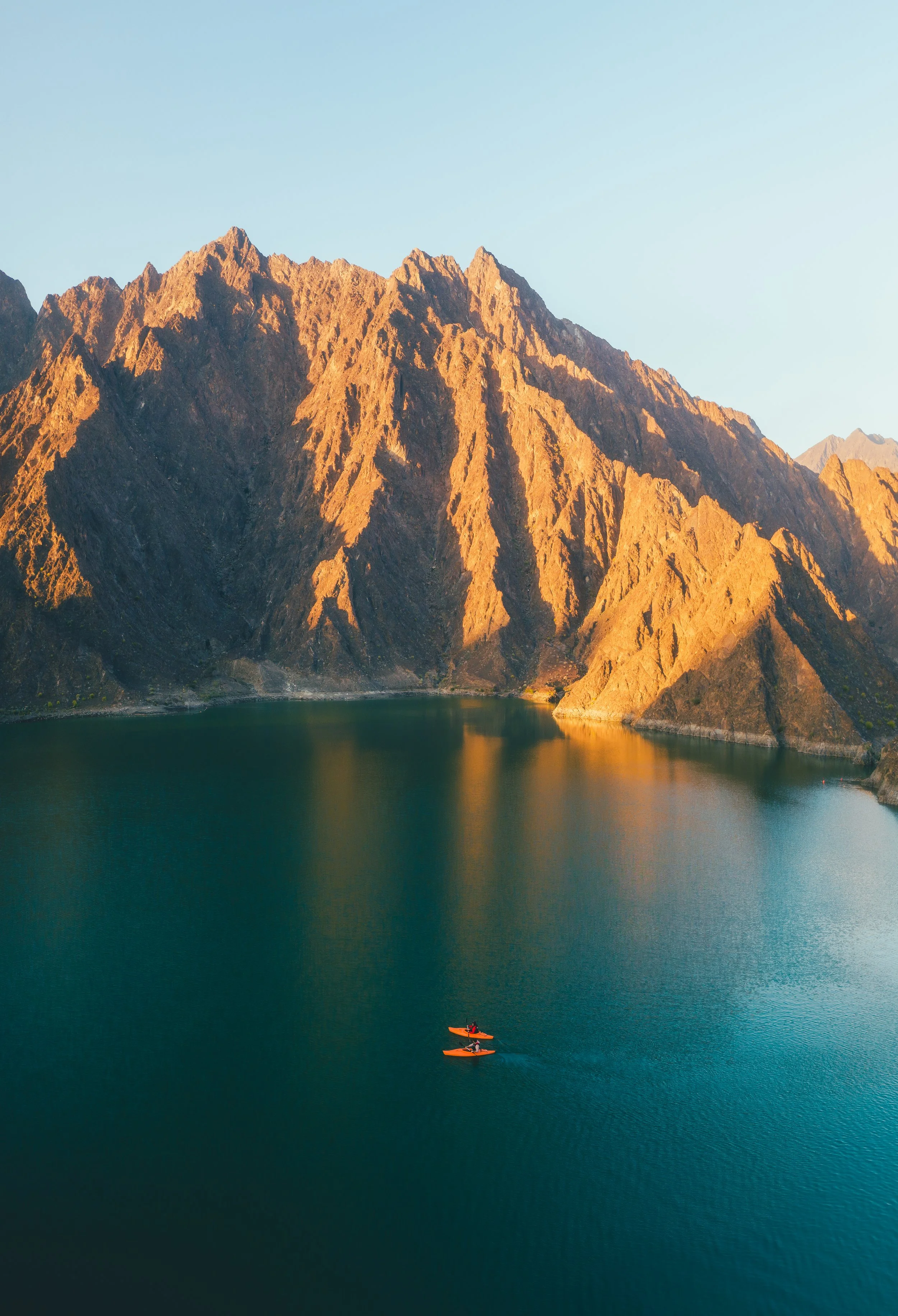 Two kayakers paddling on a calm water body near towering mountains during sunset.